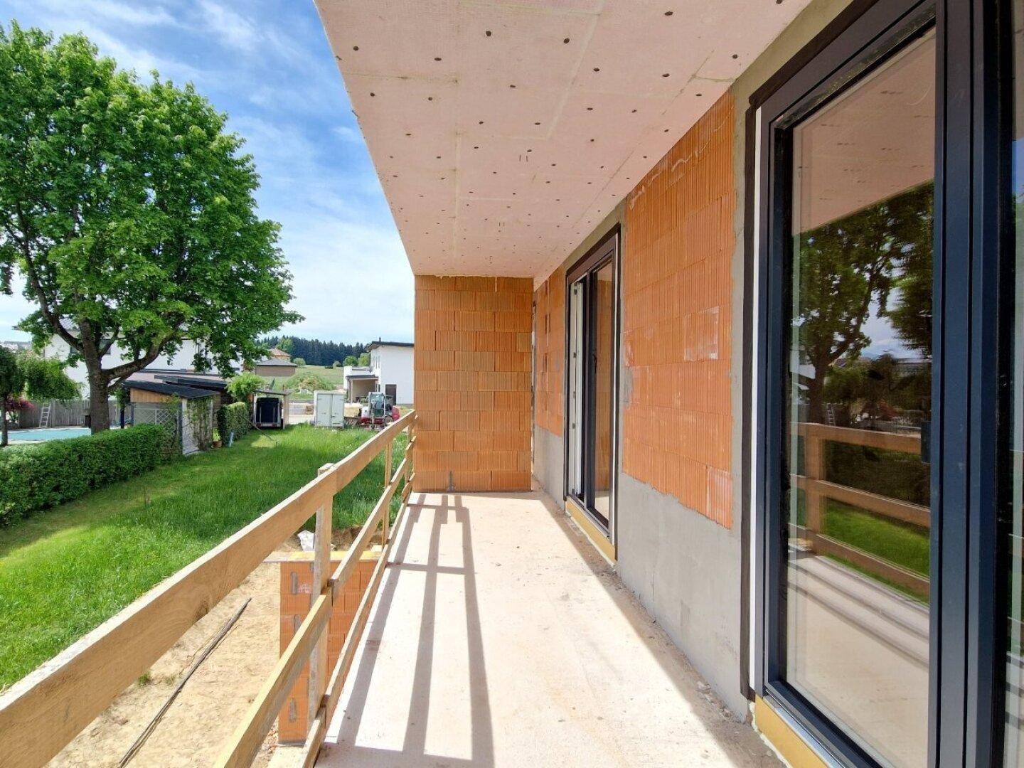 Balkon im Rohbau mit Holzgeländer und großen Fenstern, umgeben von Grün.