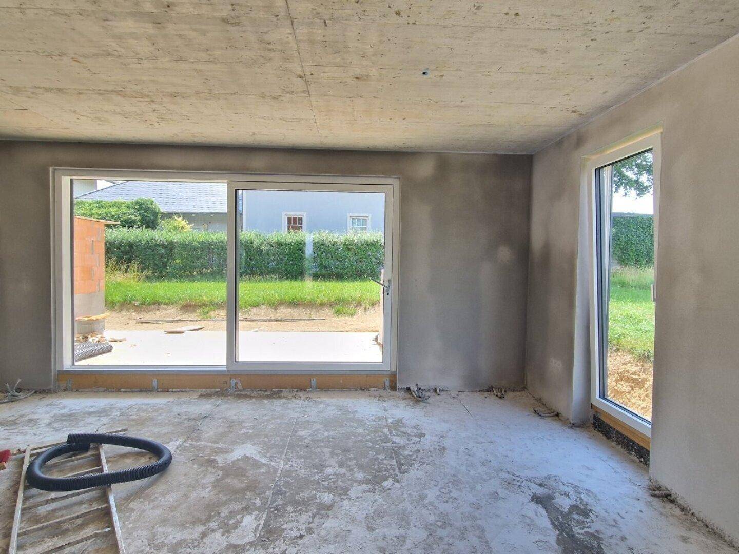Unfertiger Innenraum mit großen Fenstern und Blick ins Grüne, bereit für den Ausbau.