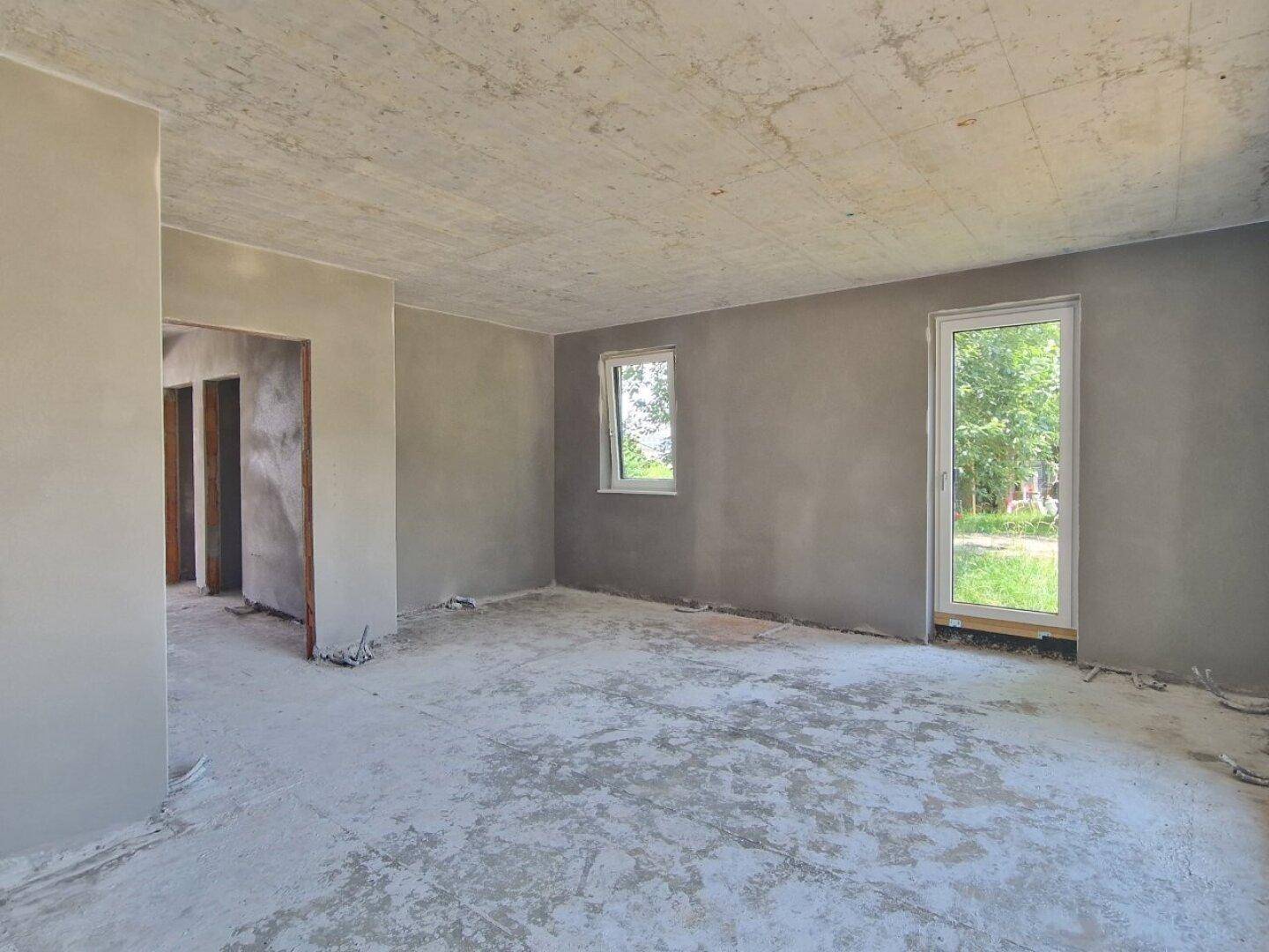 Heller Rohbau-Innenraum mit verputzten Wänden, Rohbetondecke und Blick ins Grüne durch die Fenster.
