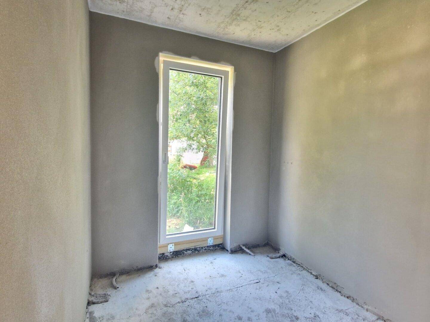 Kleiner Rohbau-Raum mit verputzten Wänden, Rohbetondecke und einem hohen Fenster mit Grünblick.