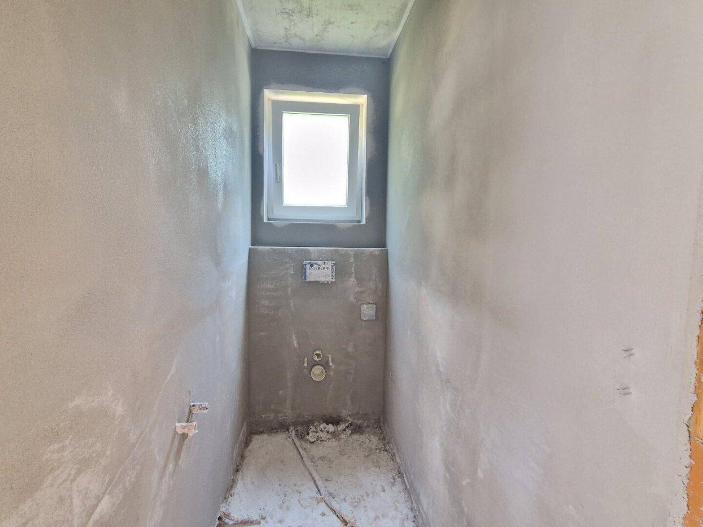 Rohbau-WC mit vorbereiteten Anschlüssen für Toilette und Waschbecken sowie einem kleinen Fenster.