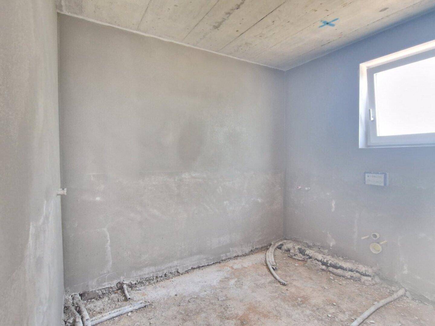 Rohbau-Badezimmer mit vorbereiteten Anschlüssen für Sanitäranlagen und einem Fenster.