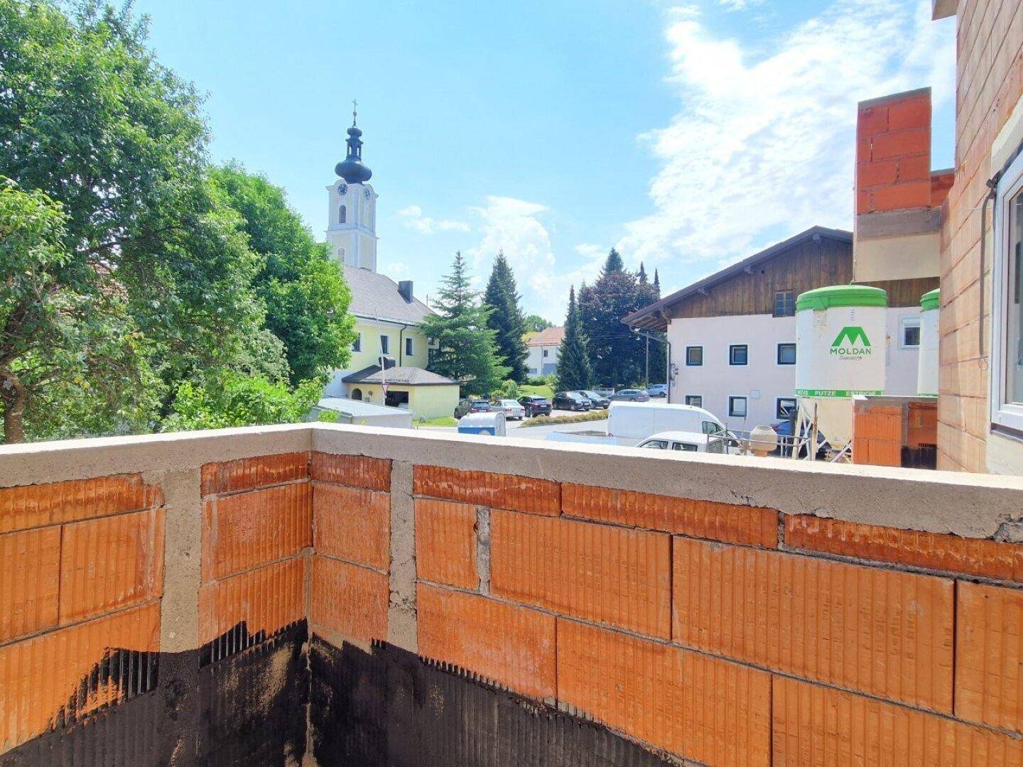 Blick von einem Rohbaubalkon auf die umliegende Bebauung und einen Kirchturm.