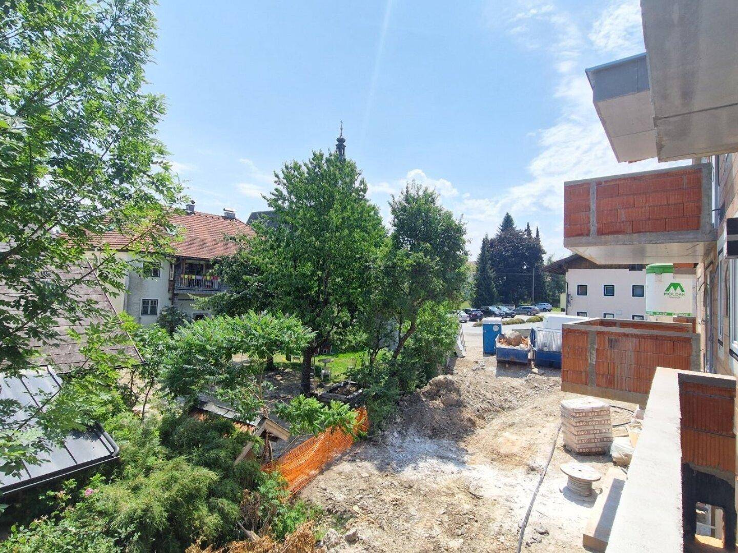 Blick von einem Balkon auf die Baustelle, umliegende Bäume und Dächer in der Ferne.
