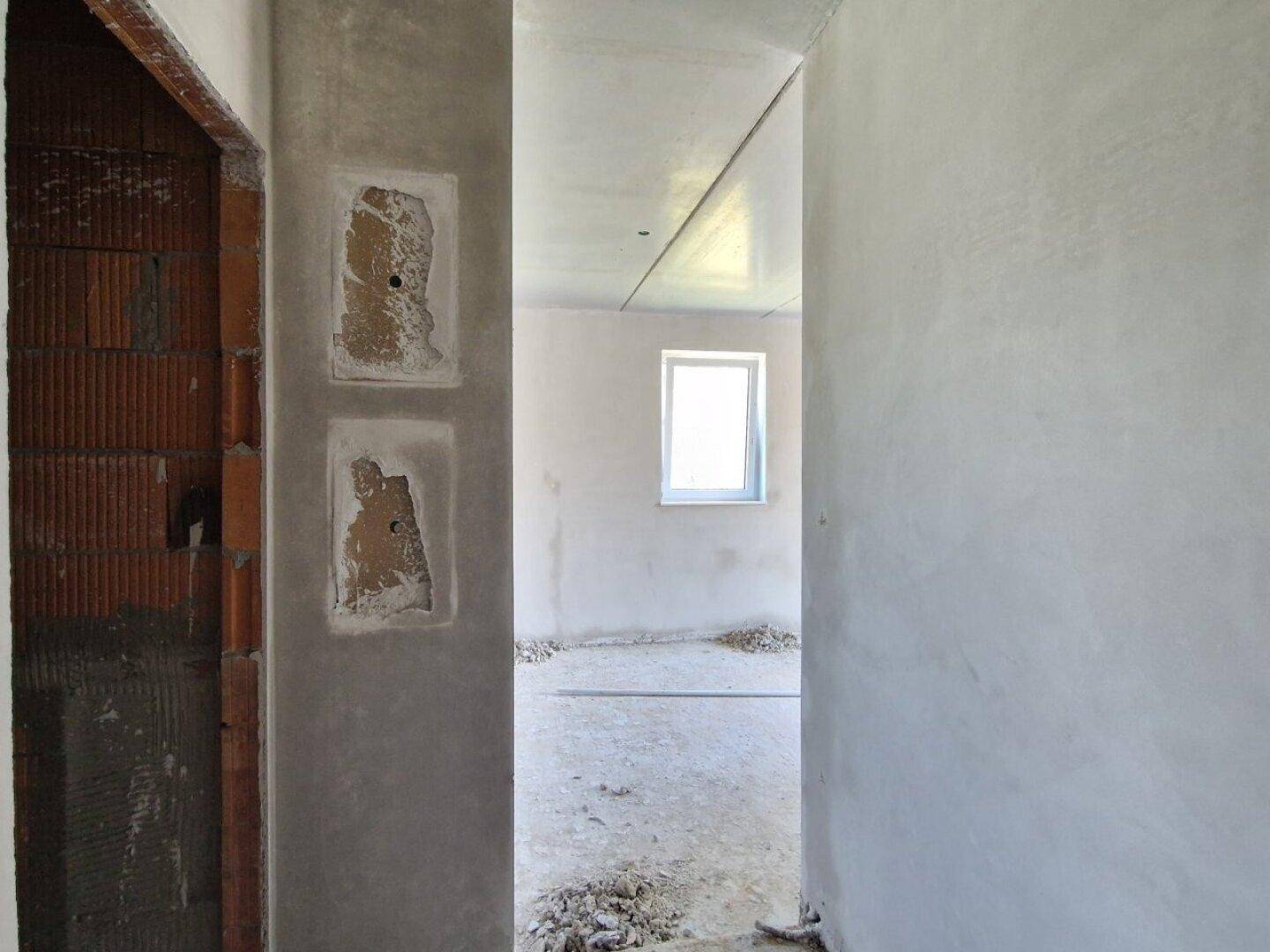 Blick durch einen Türrahmen in einen weiteren unfertigen Raum mit Fenster im Rohbauzustand.