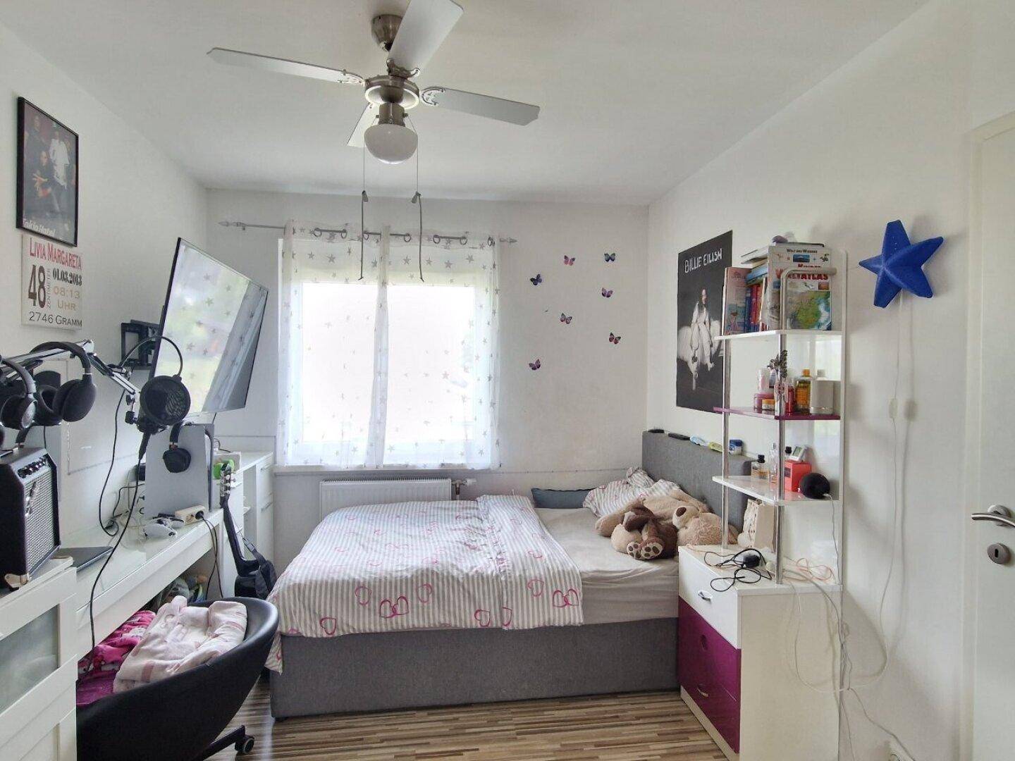 Helles Schlafzimmer mit Bett, Schreibtisch, Deckenventilator und Holzboden.