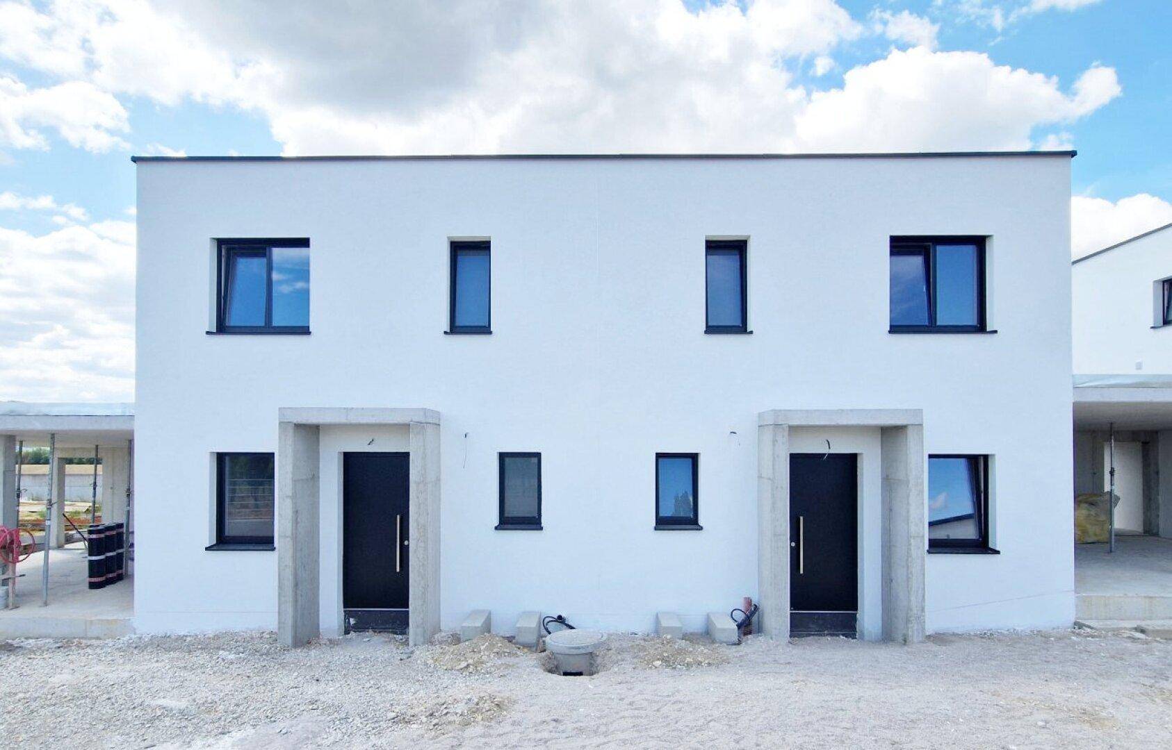 Frontansicht eines modernen Neubaus mit weißer Fassade und schwarzen Fensterrahmen im Rohbau.