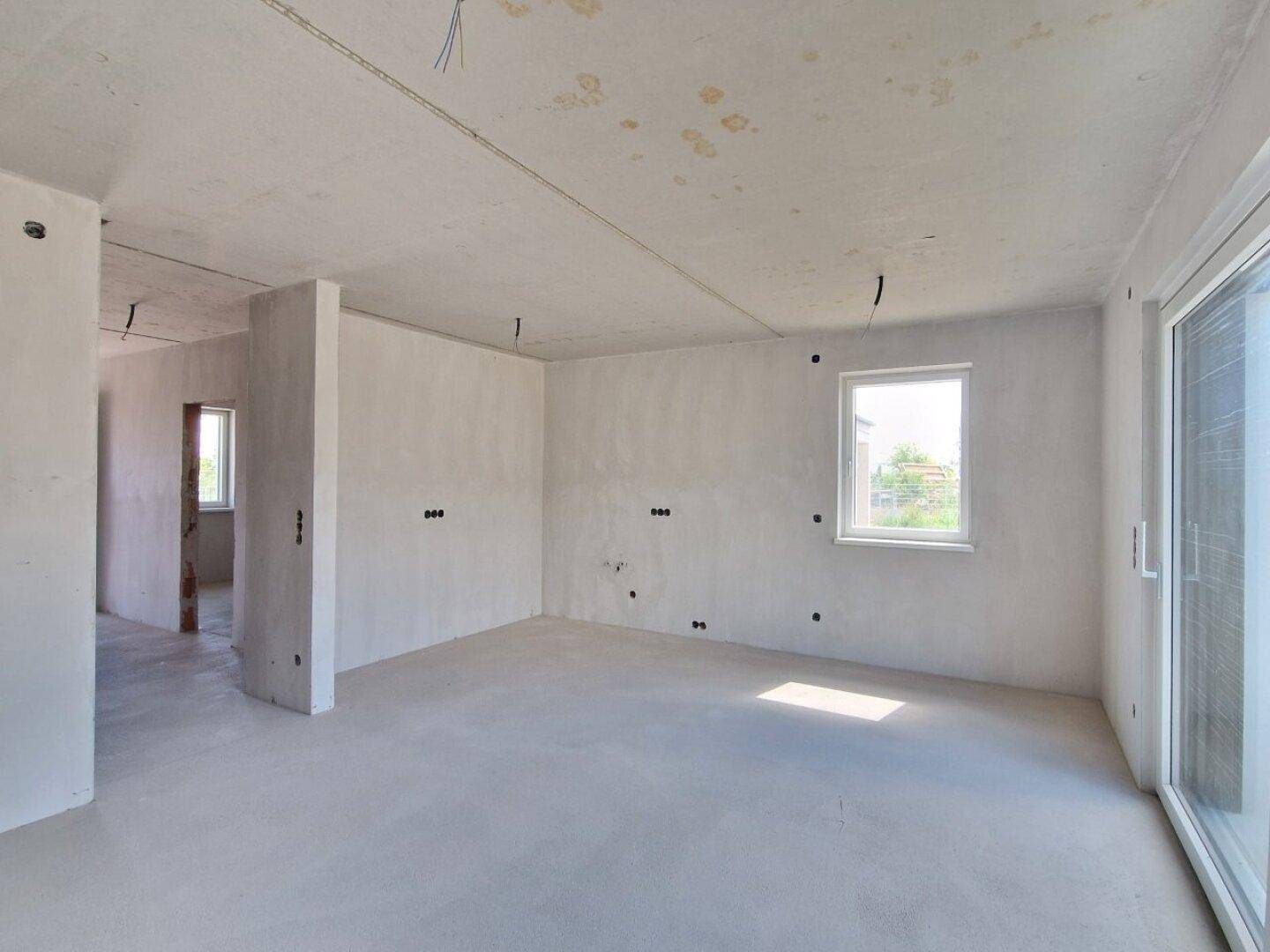 Geräumiger Wohnbereich im Rohbau mit viel Tageslicht und unfertigen Wänden und Boden.