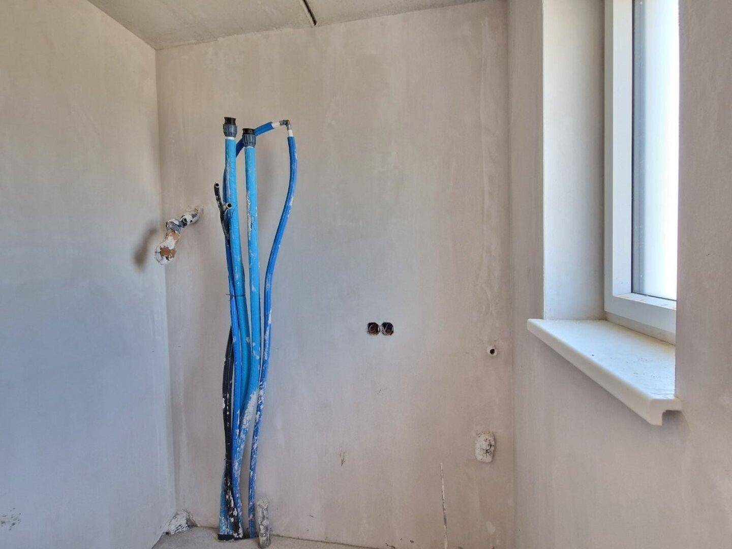 Badezimmer im Rohbau mit sichtbaren Wasserleitungen und einem Fenster für Tageslicht.