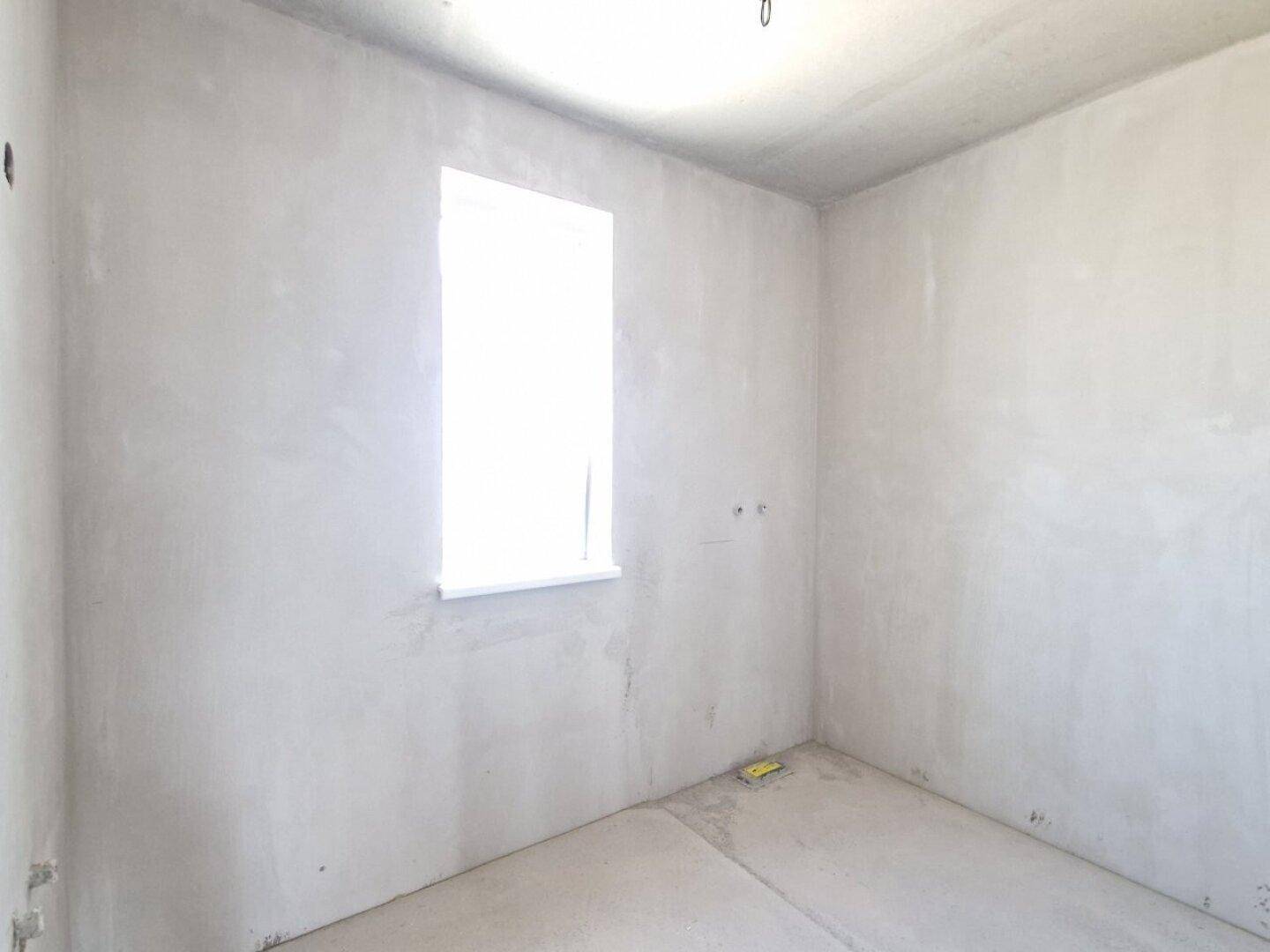 Helles Schlafzimmer im Rohbau mit einem Fenster und unfertigen Oberflächen und Boden.