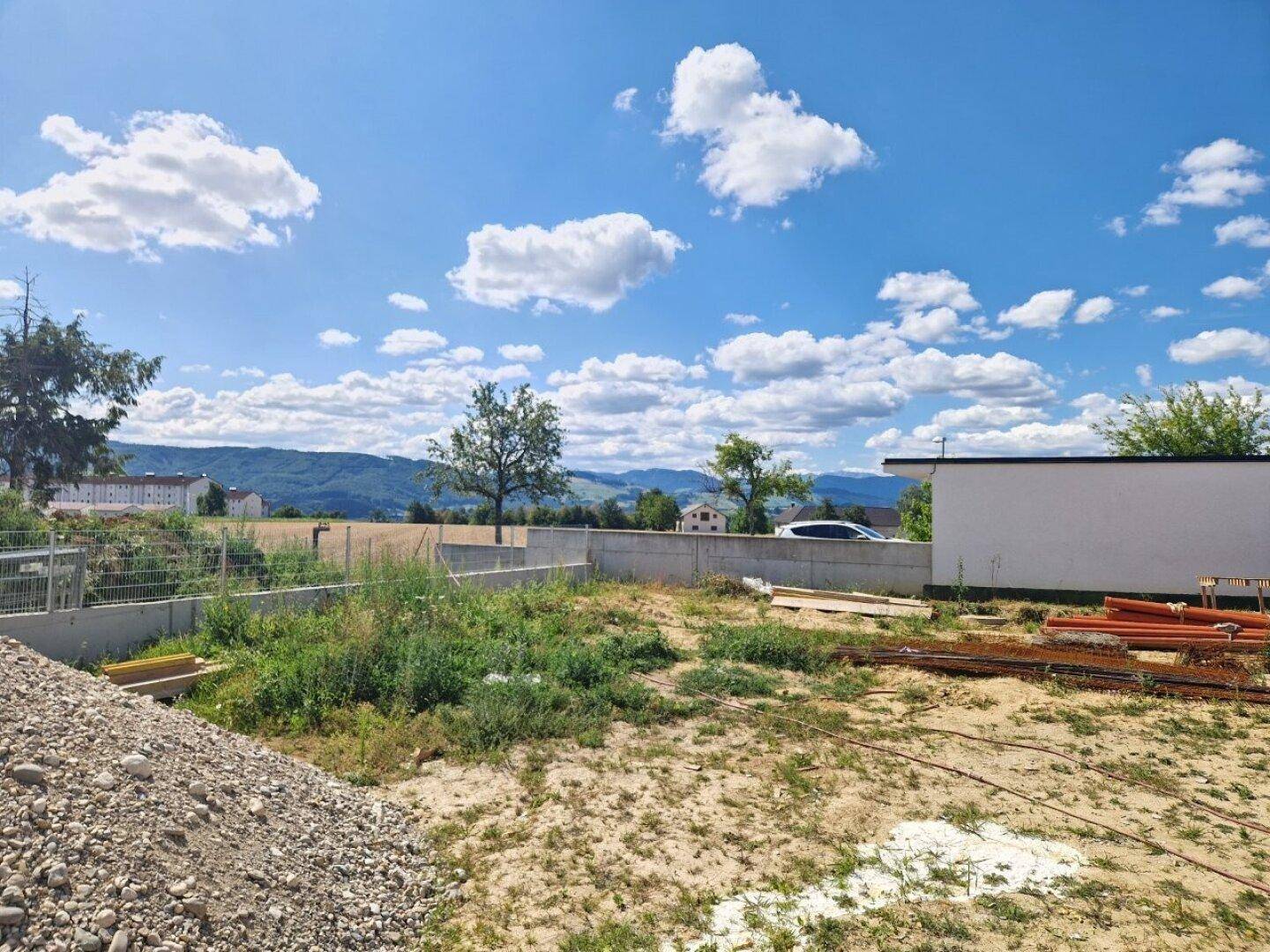 Baustelle mit Erdhaufen und Baumaterialien, umgeben von Natur und Bergblick unter blauem Himmel.