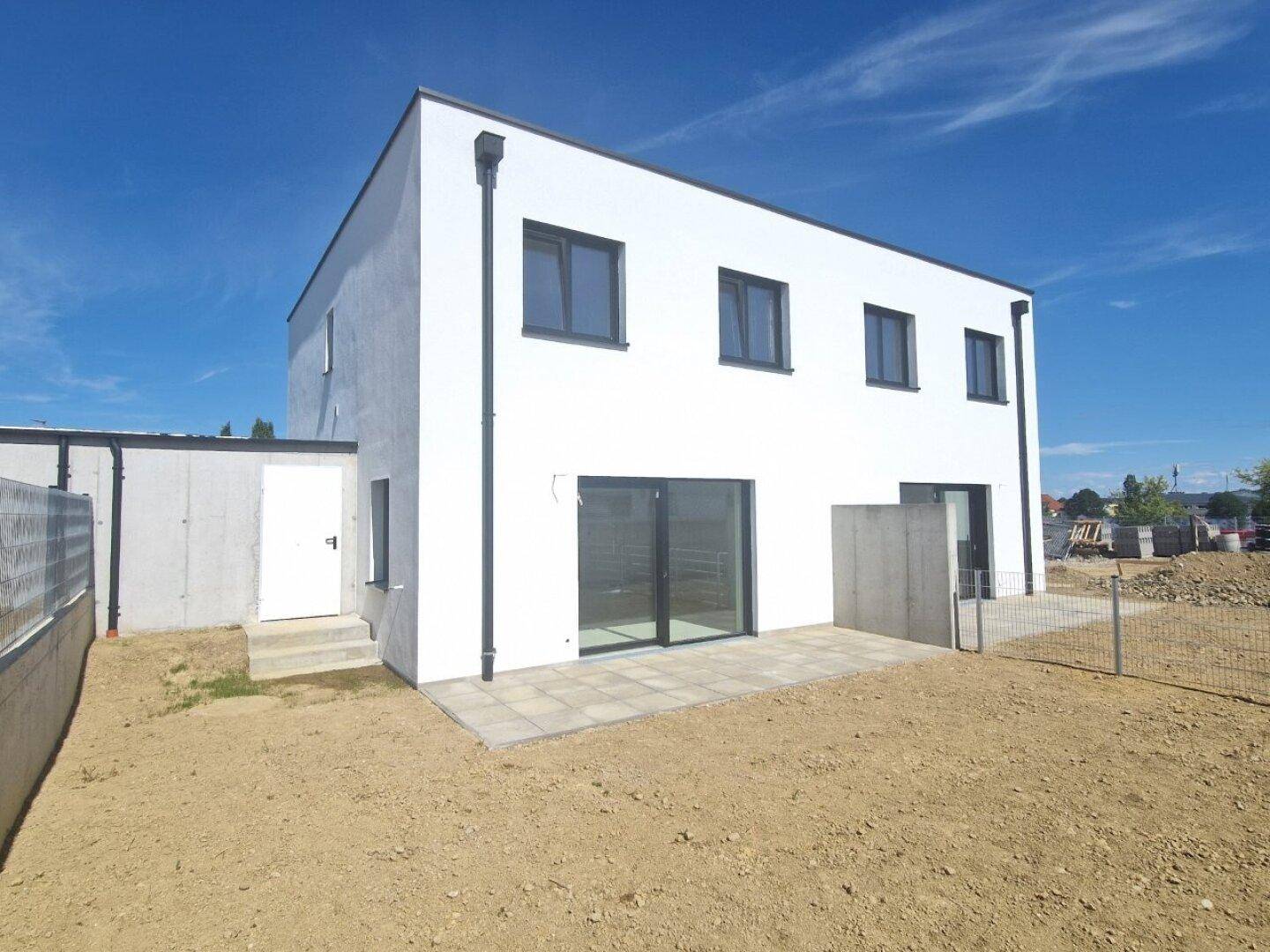 Rückseite eines Neubaus mit kleiner gepflasterter Terrasse und ungestaltetem Gartenbereich.