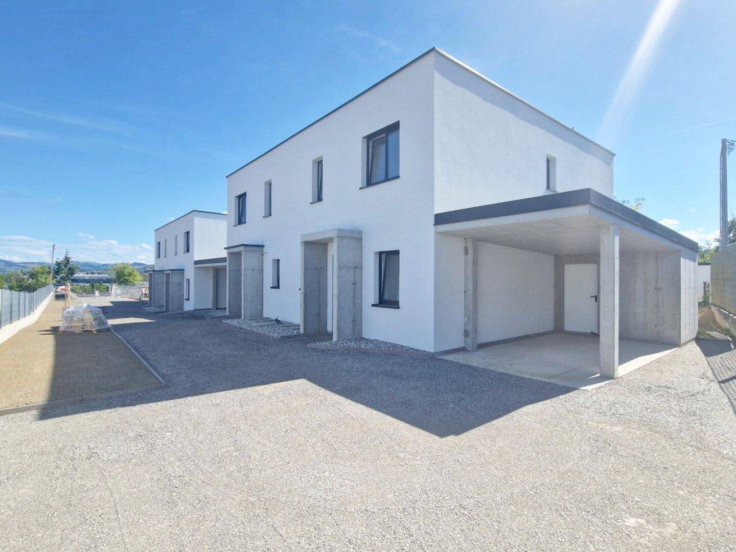 Moderne Reihenhäuser im Neubau mit Carports und Schotterzufahrt unter strahlend blauem Himmel.