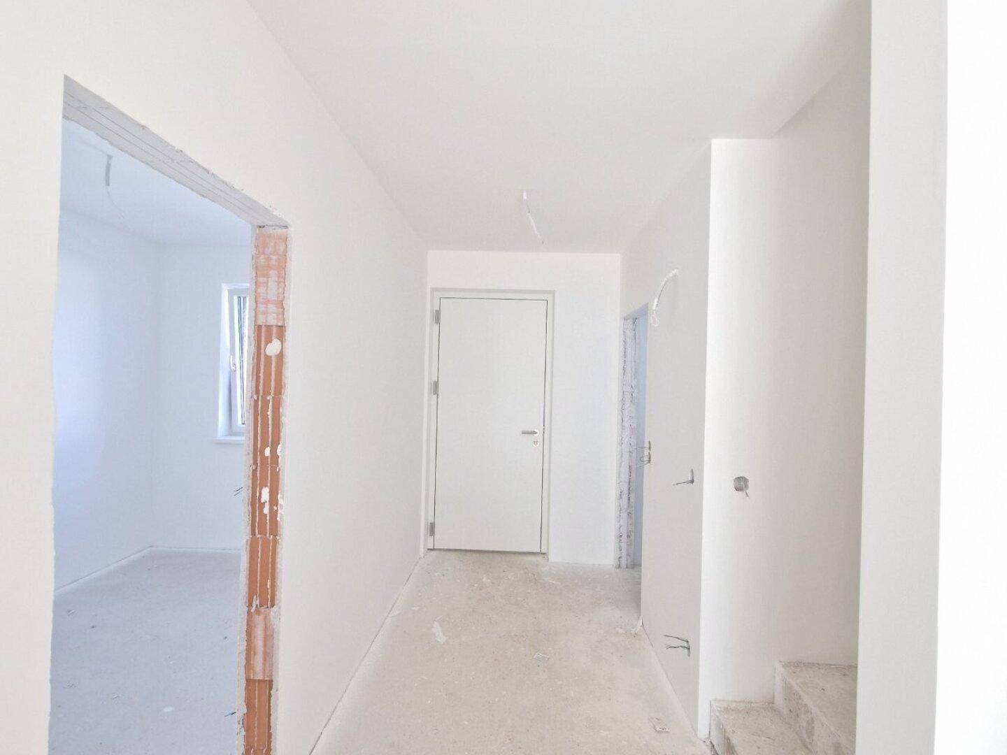 Flur im Rohbau mit weißer Tür und offenem Durchgang zu einem weiteren Zimmer.
