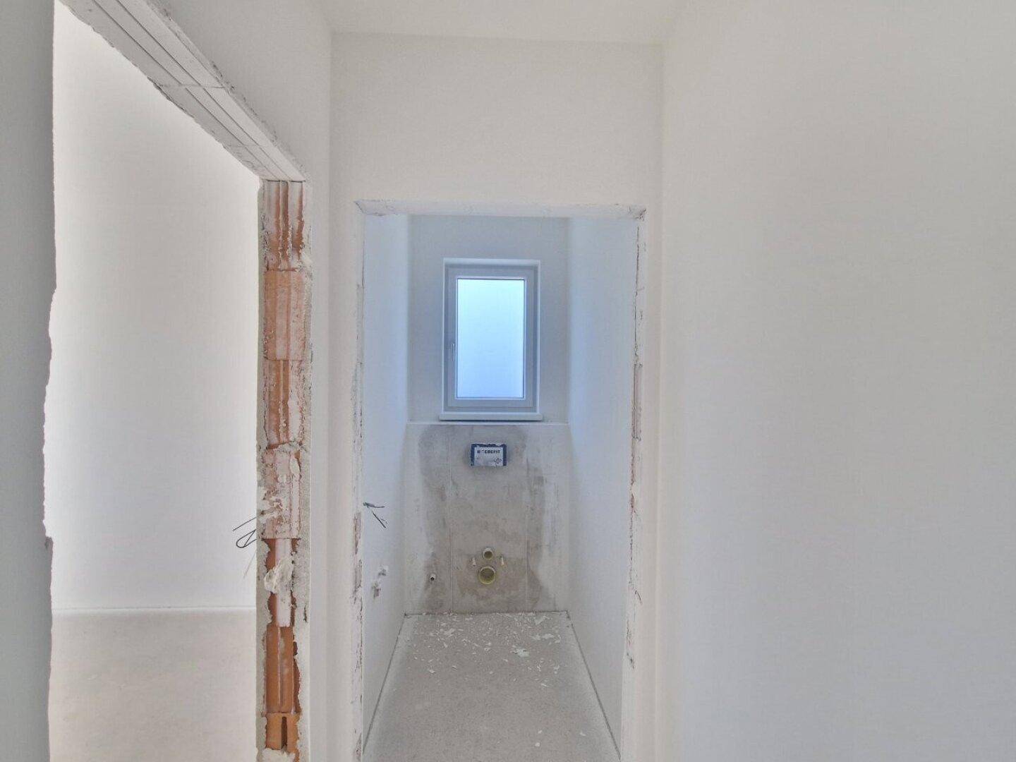 Kleiner Raum im Rohbau mit Fenster und vorbereiteten Anschlüssen für ein Wand-WC.
