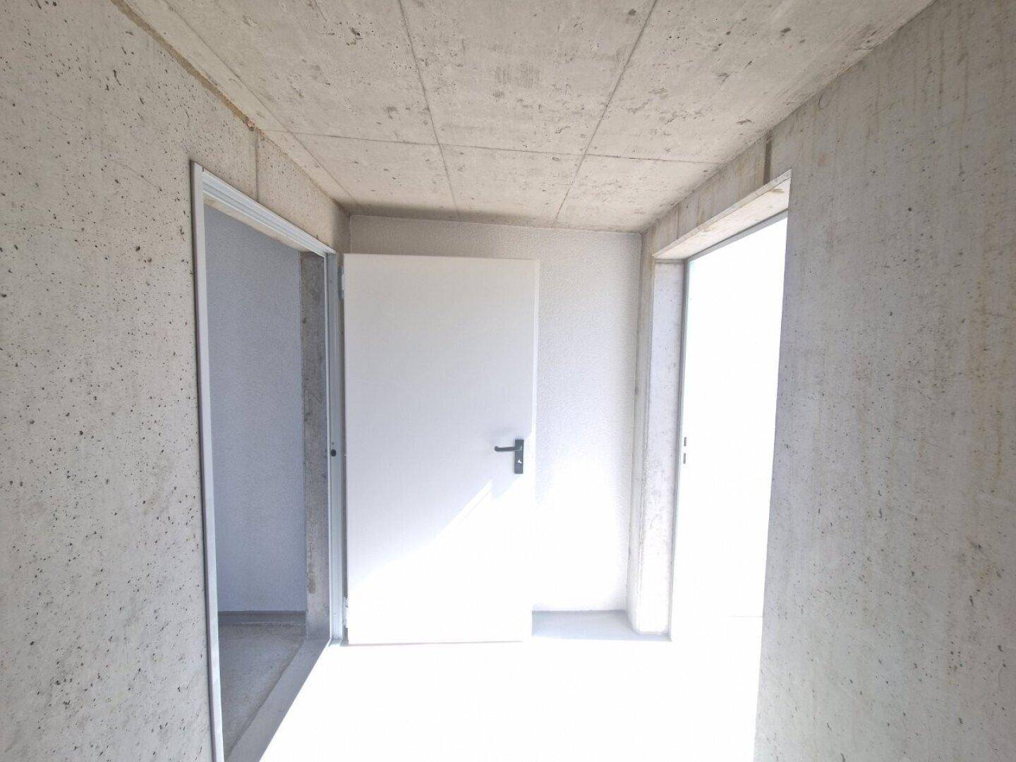 Lagerraum im Rohbau mit Sichtbetonwänden und zwei weißen Türen, hell durch Tageslicht.