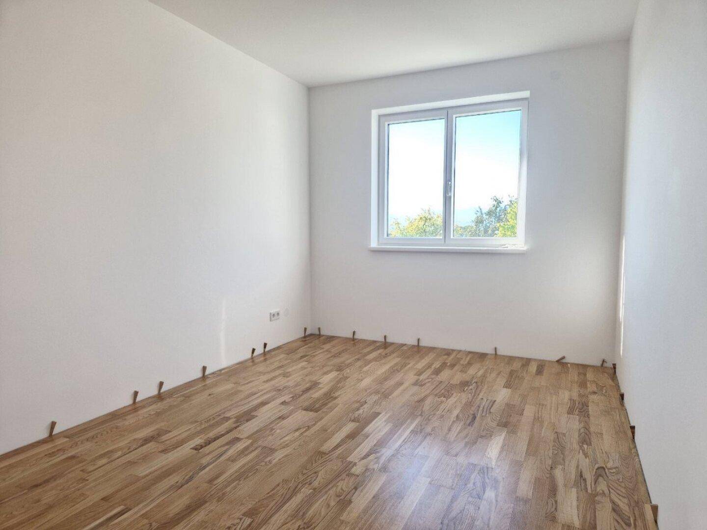 Leeres Zimmer im Neubau mit Parkettboden und einem großen Fenster für viel Tageslicht.