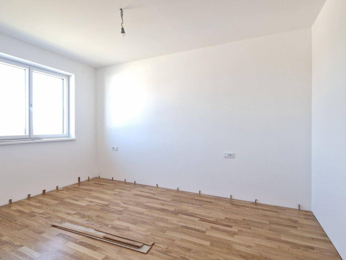 Helles Zimmer im Neubau mit frisch verlegtem Parkettboden und einem Fenster.