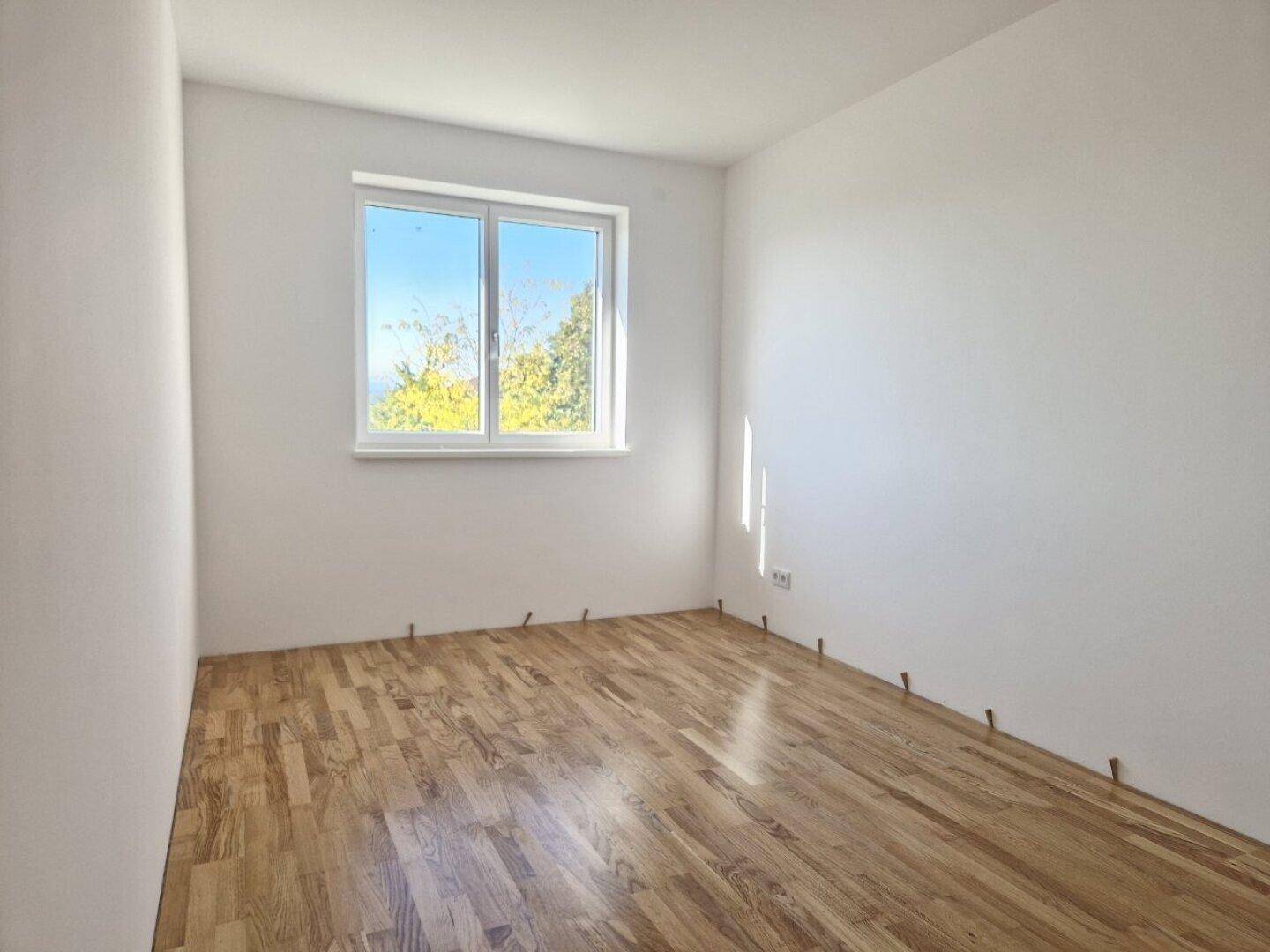 Unmöbliertes Zimmer im Neubau mit Holzboden und Blick ins Grüne durch das Fenster.
