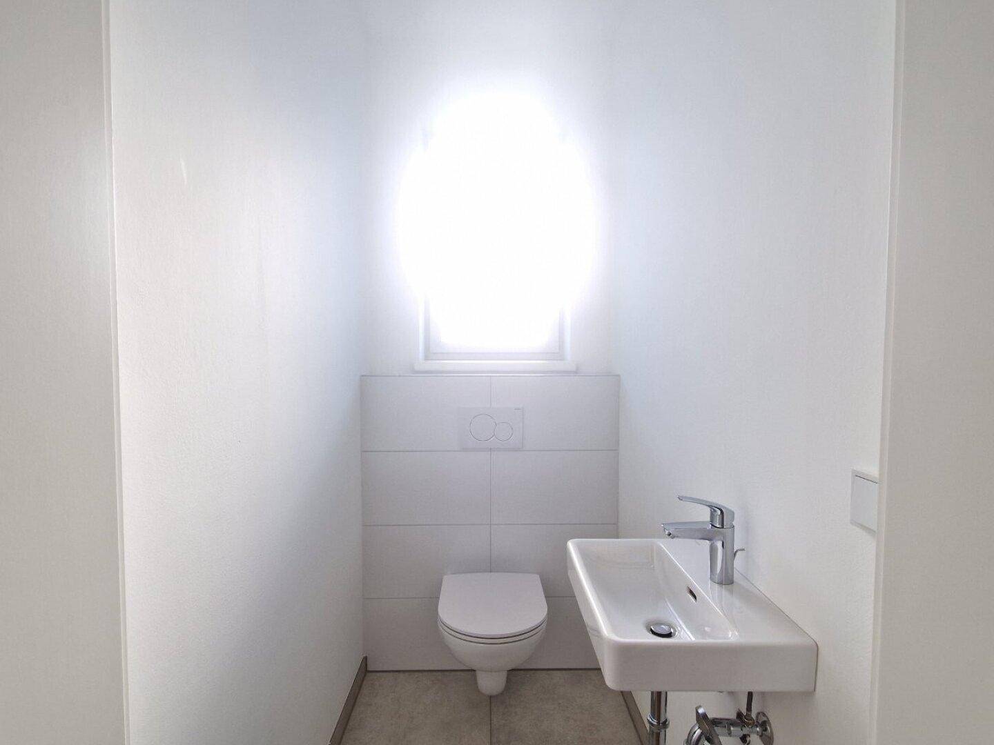 Helles Gäste-WC mit wandhängendem WC, kleinem Waschbecken und Fenster.