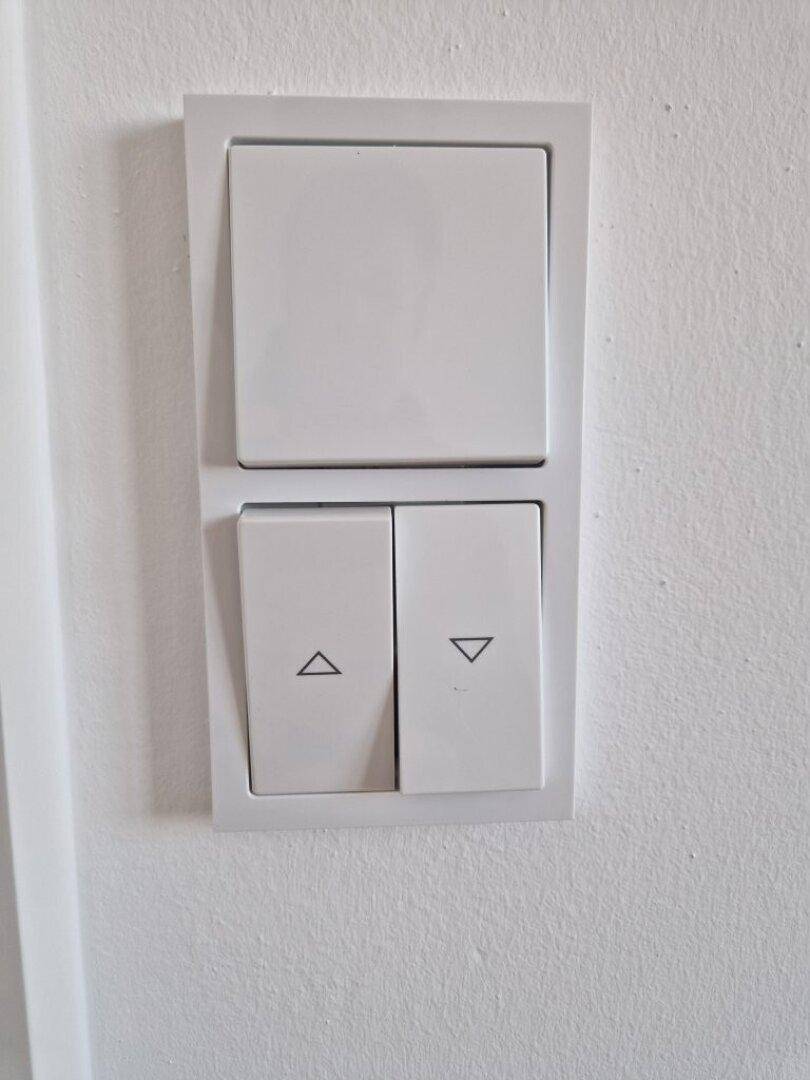 Detailaufnahme von weißen Lichtschaltern und Rollladensteuerung an einer hellen Wand.