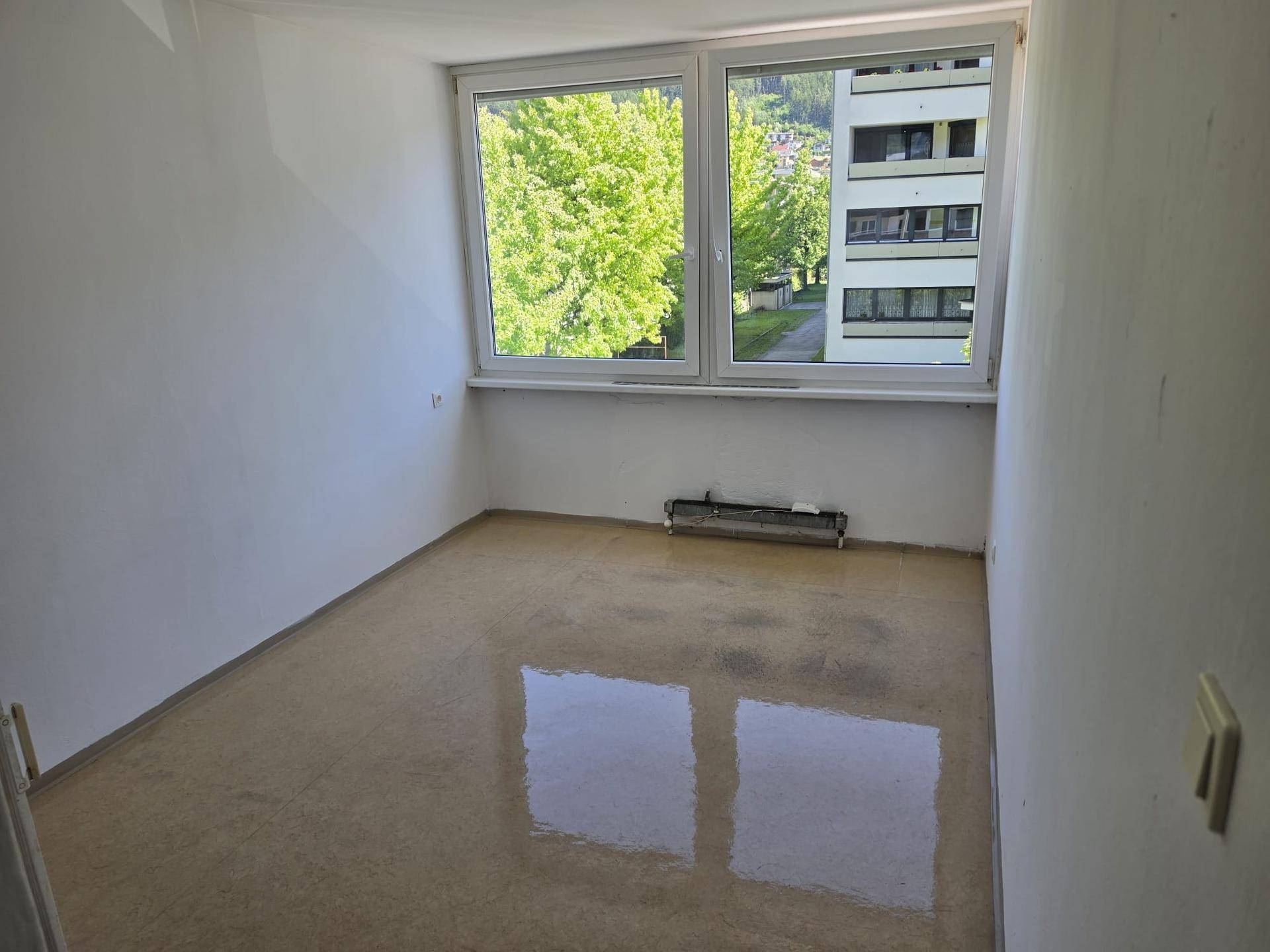 Leeres Zimmer mit großen Fenstern, Blick ins Grüne und auf ein Gebäude, bereit für neue Bodenbeläge.