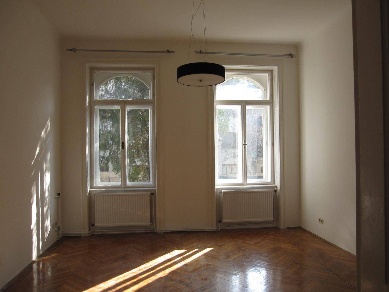Lichtdurchfluteter Raum mit zwei hohen Fenstern, Heizkörpern und klassischem Fischgrätparkettboden.