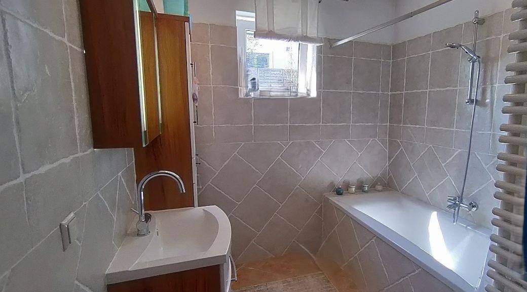 Badezimmer mit hellgrauen Fliesen, Badewanne, Einzelwaschbecken und einem Fenster für Tageslicht.