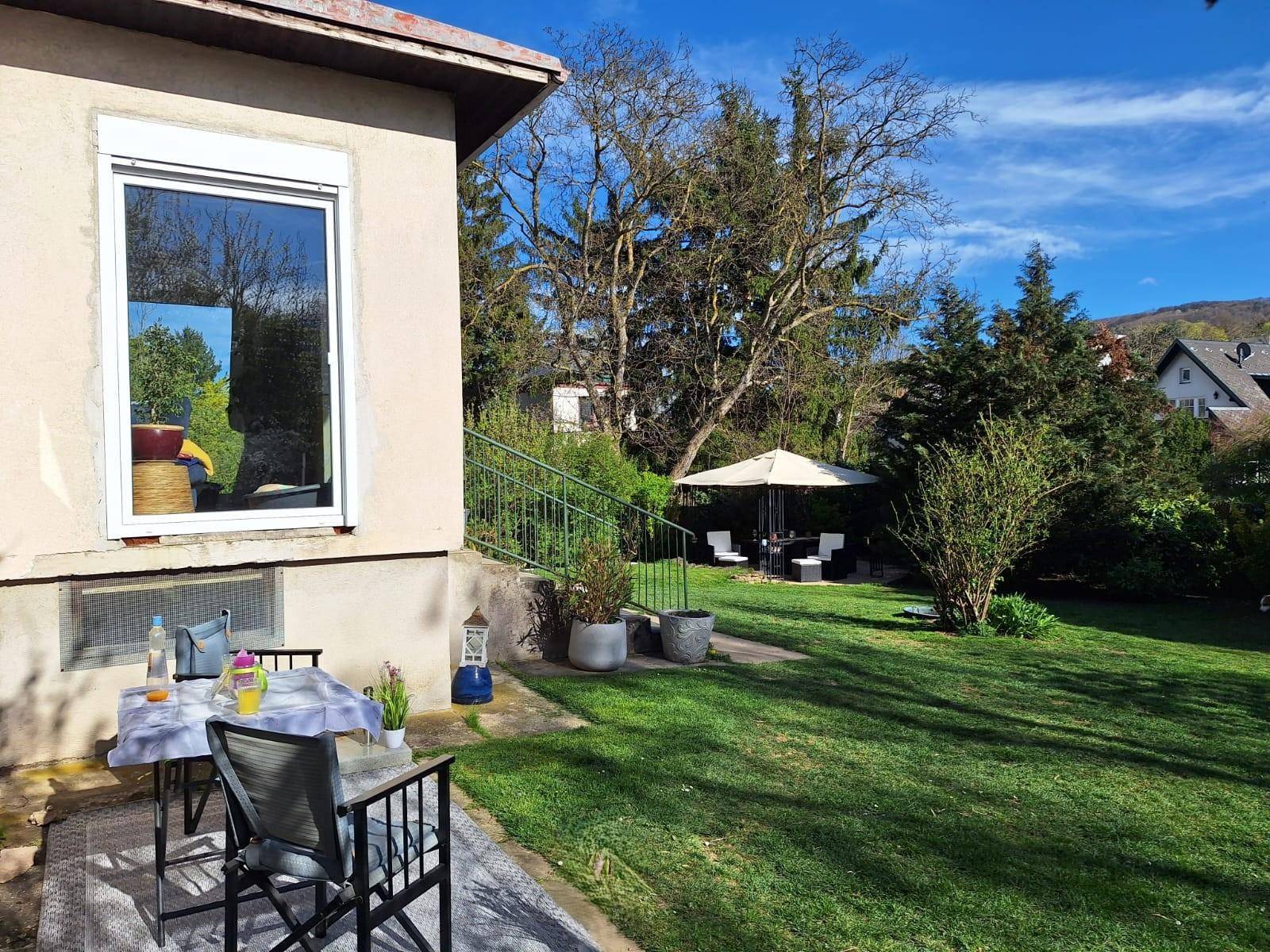 Kleine gepflasterte Terrasse am Haus mit Gartentisch und Stühlen, umgeben von Grünflächen.