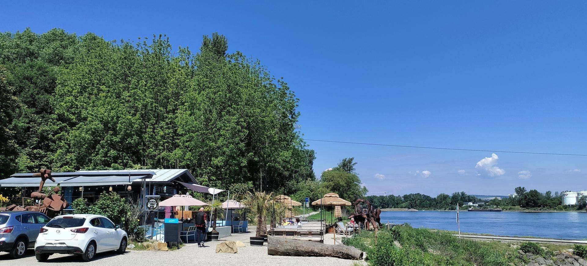 Flussufer mit einem Restaurantbereich, Parkplätzen und üppiger Vegetation im Hintergrund.