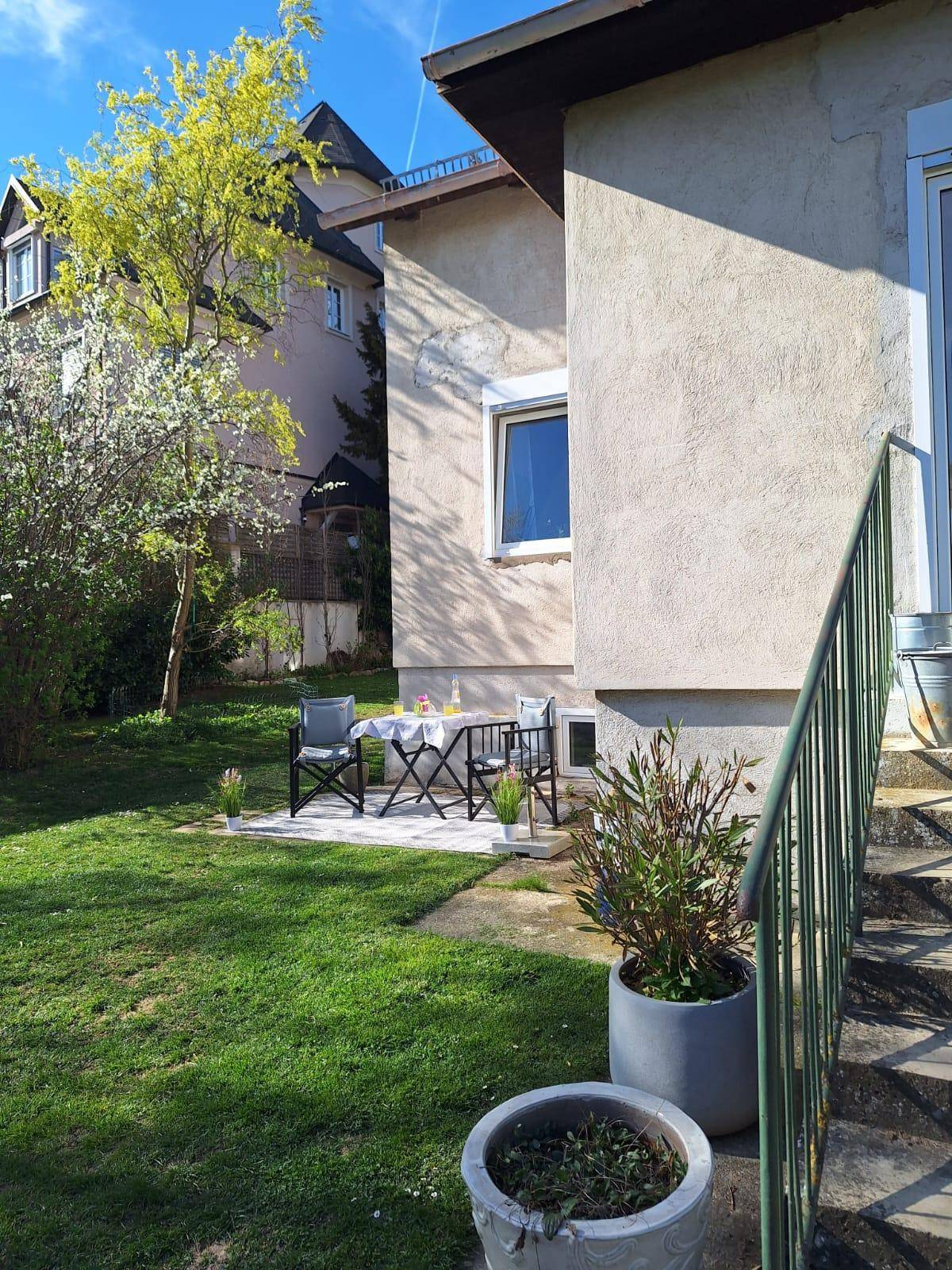Kleine Terrasse am Haus mit Gartentisch und Stühlen, umgeben von Rasen und Sträuchern.