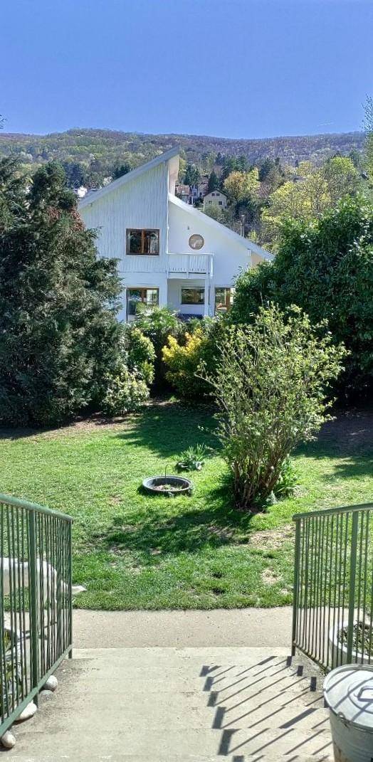 Blick auf ein weißes Haus mit Satteldach, umgeben von einem großen grünen Garten und Bäumen.