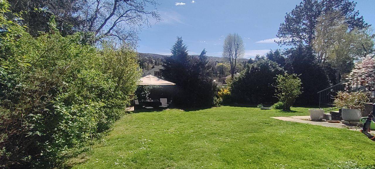 Weitläufiger grüner Garten mit Rasenfläche, hohen Bäumen und einem schattigen Sitzbereich im Hintergrund.