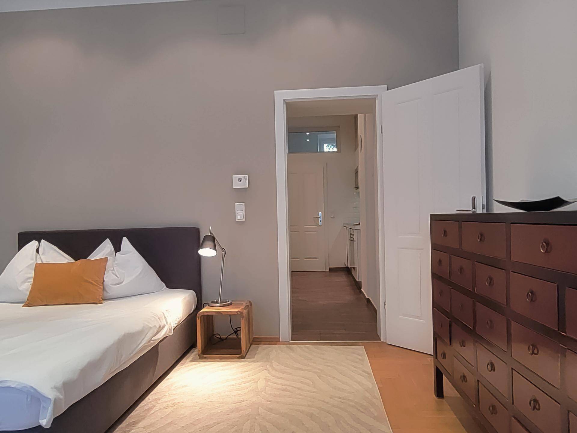 Schlafzimmer mit Doppelbett, Nachttisch und Blick in die angrenzende Küche.
