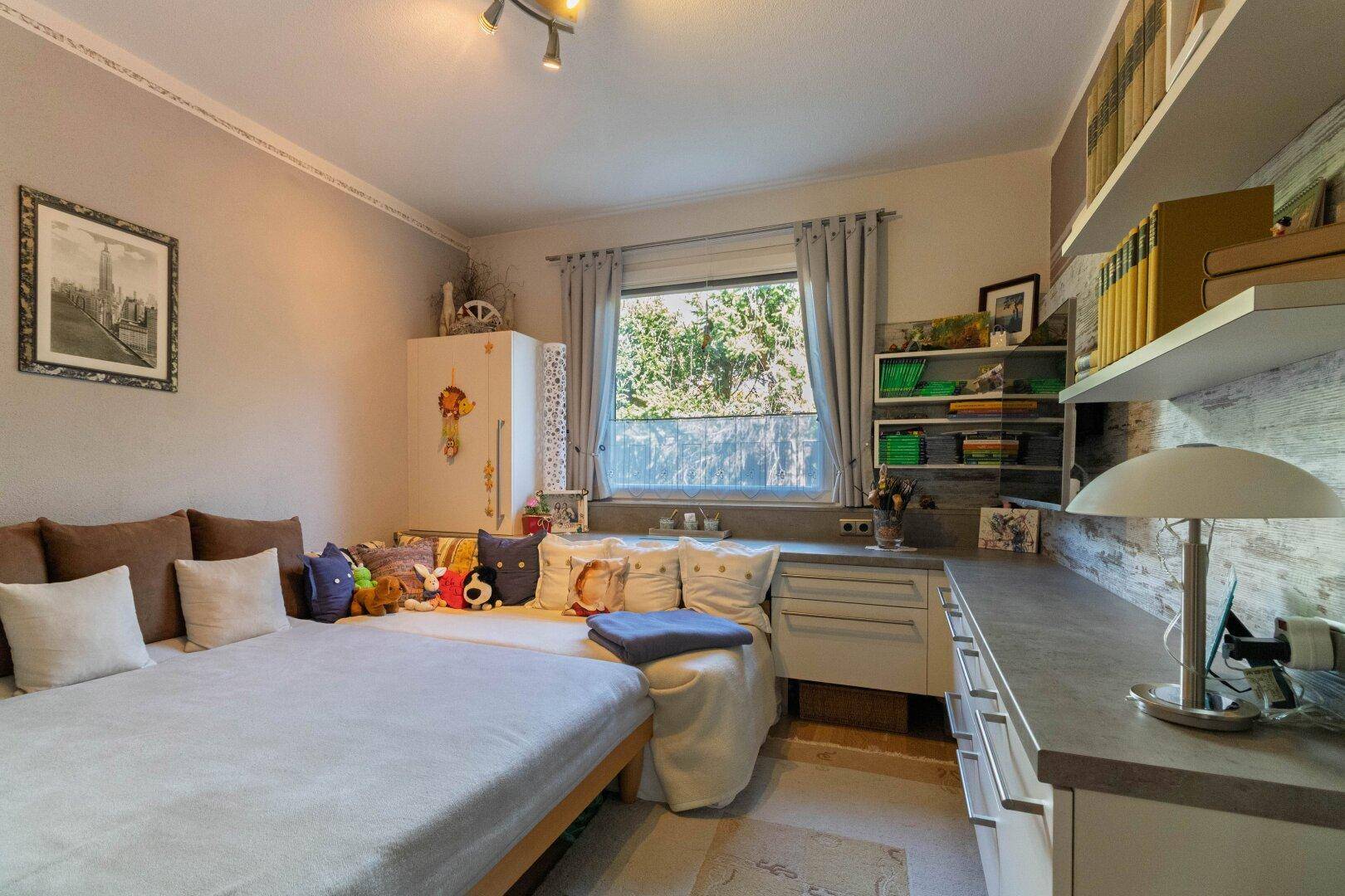 Helles Kinder- oder Gästezimmer mit Bett, Schreibtisch und Fenster zum Grünen.