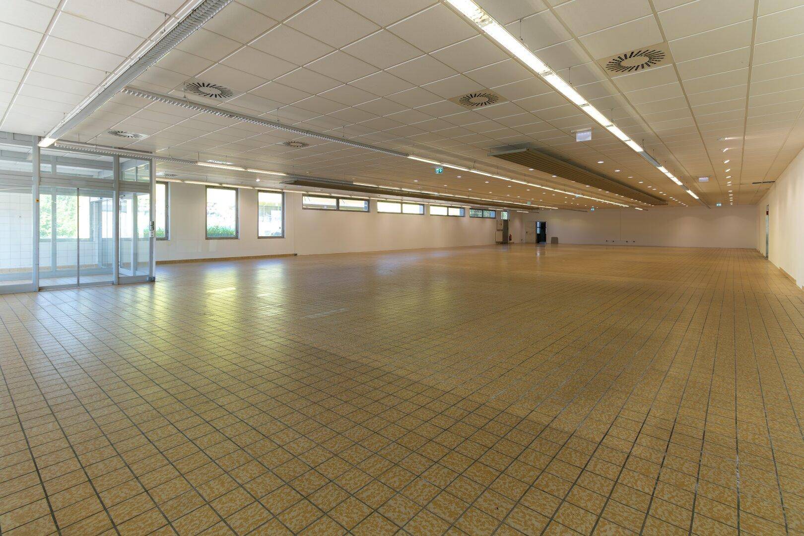 Großer, leerer Gewerberaum mit Fliesenboden, abgehängter Decke und vielen Fenstern für natürliches Licht.