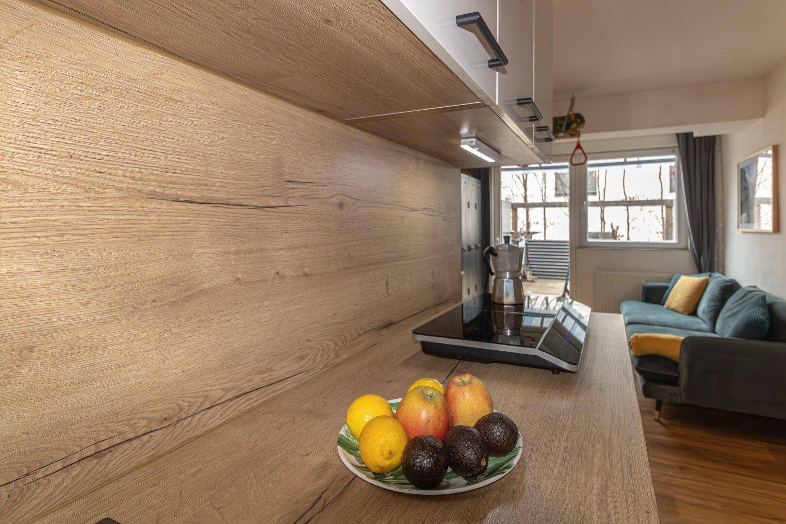 Offener Wohnbereich mit Küchenzeile, Holz-Arbeitsplatte und Blick auf das Sofa und die Fenster.
