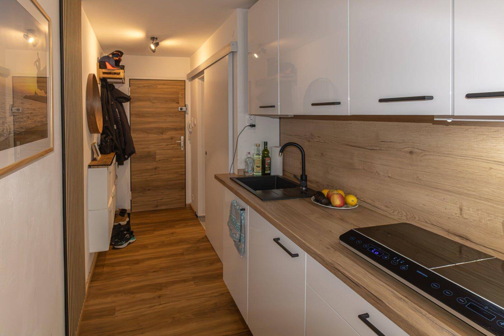 Moderne Küchenzeile mit weißen Hochglanzschränken, Holz-Arbeitsplatte und Induktionskochfeld im Eingangsbereich.