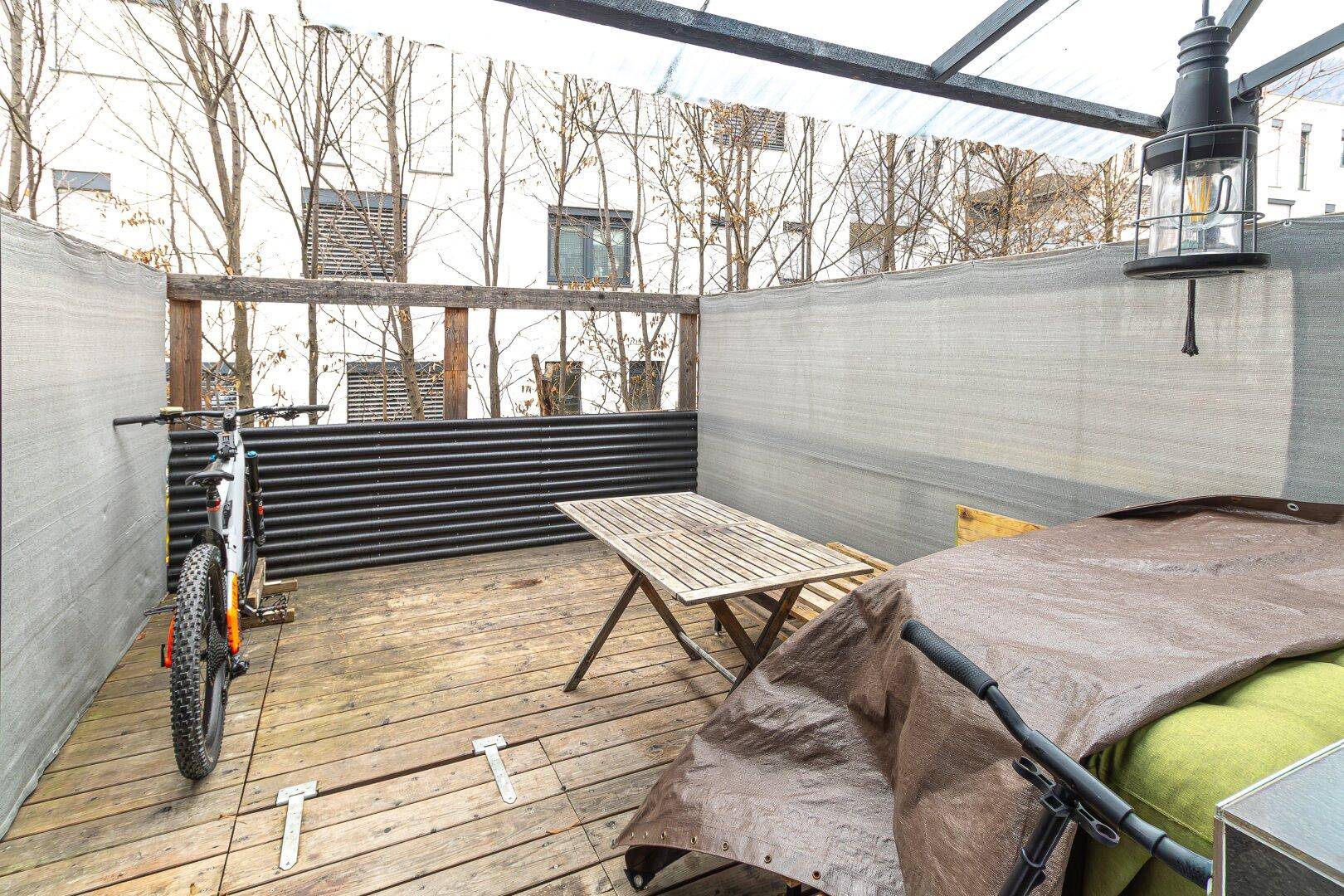 Überdachte Terrasse mit Holzboden, einem Tisch und Platz für Fahrräder, umgeben von Gebäuden.