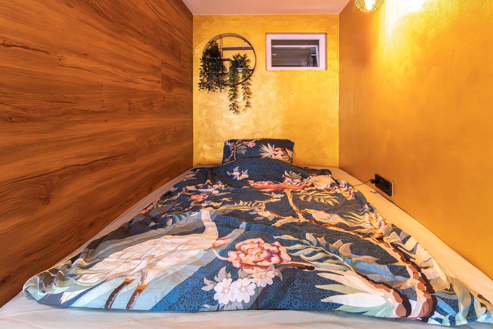 Gemütliches Schlafzimmer mit Holzwandverkleidung, gelber Wand und einem Bett mit farbenfroher Bettwäsche.