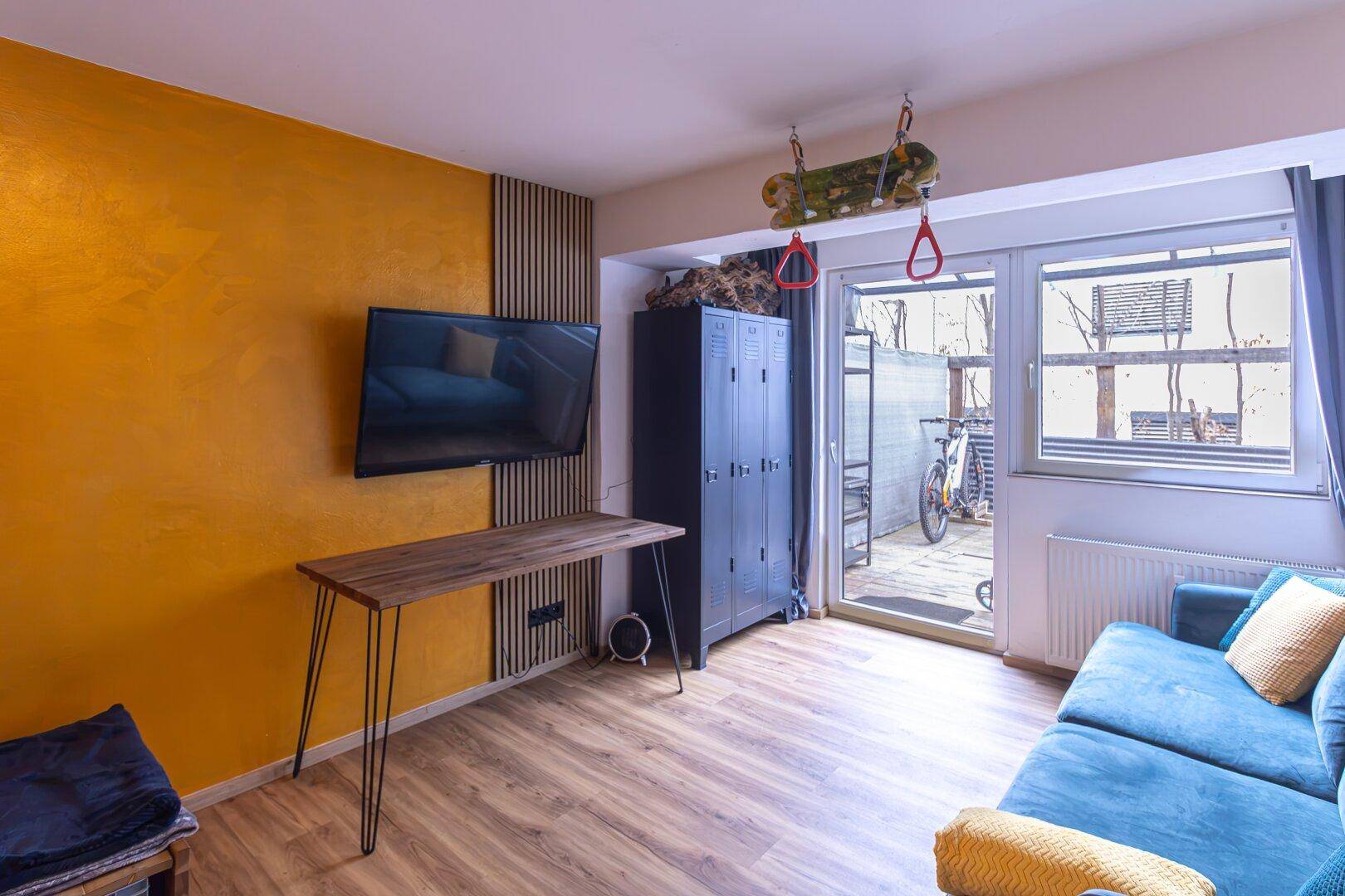 Helles Wohnzimmer mit gelber Akzentwand, Sofa, Fernseher und Zugang zu einem Außenbereich.