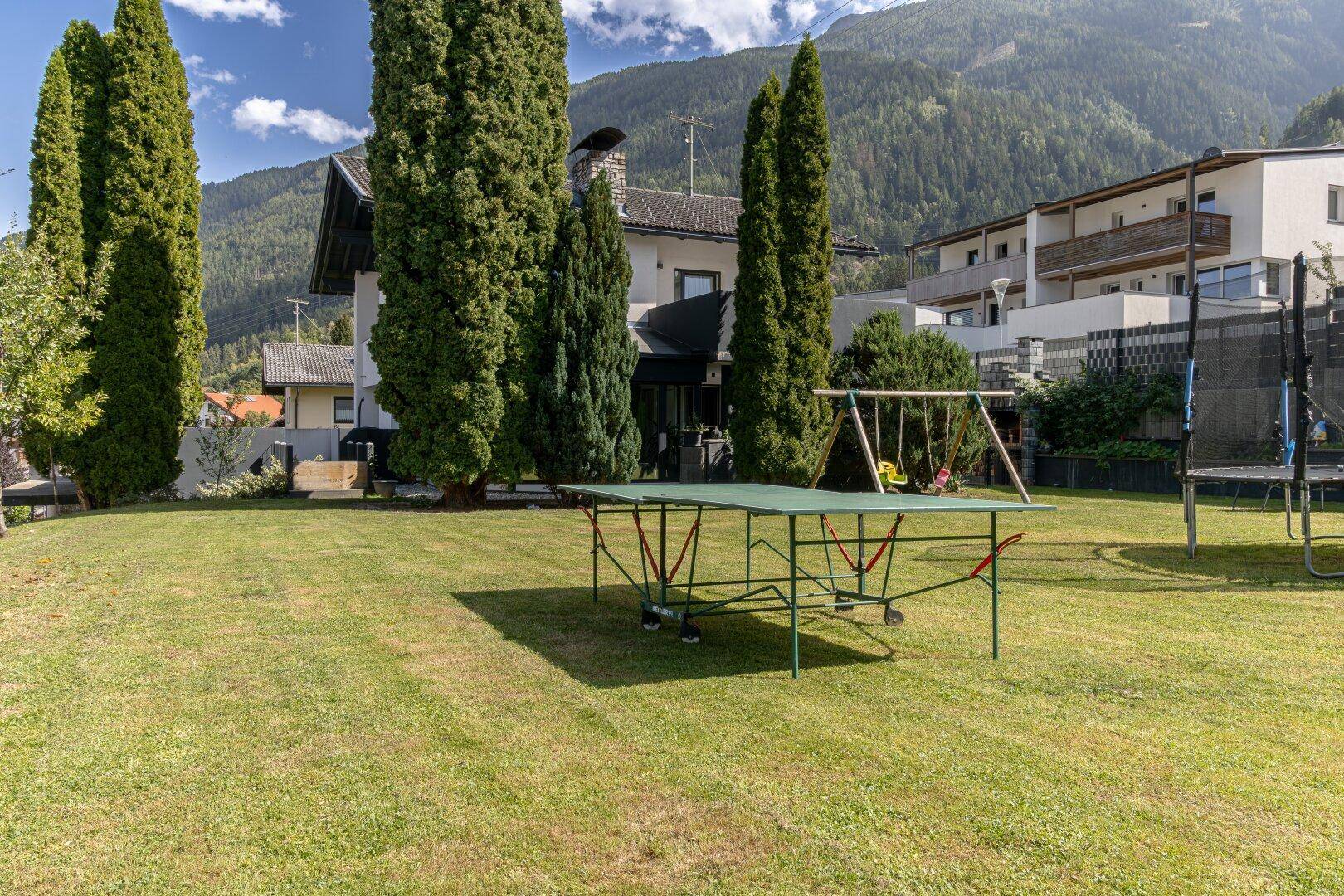 Großer, gepflegter Garten mit Tischtennisplatte, Schaukel und hohen Bäumen, ideal für Freizeitaktivitäten.