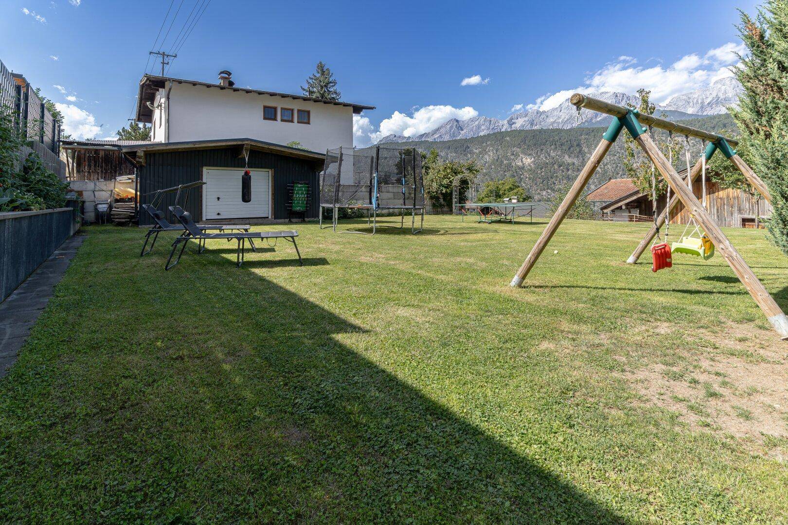 Weitläufiger Garten mit Schaukel, Trampolin und einem Nebengebäude, umgeben von Bergen.