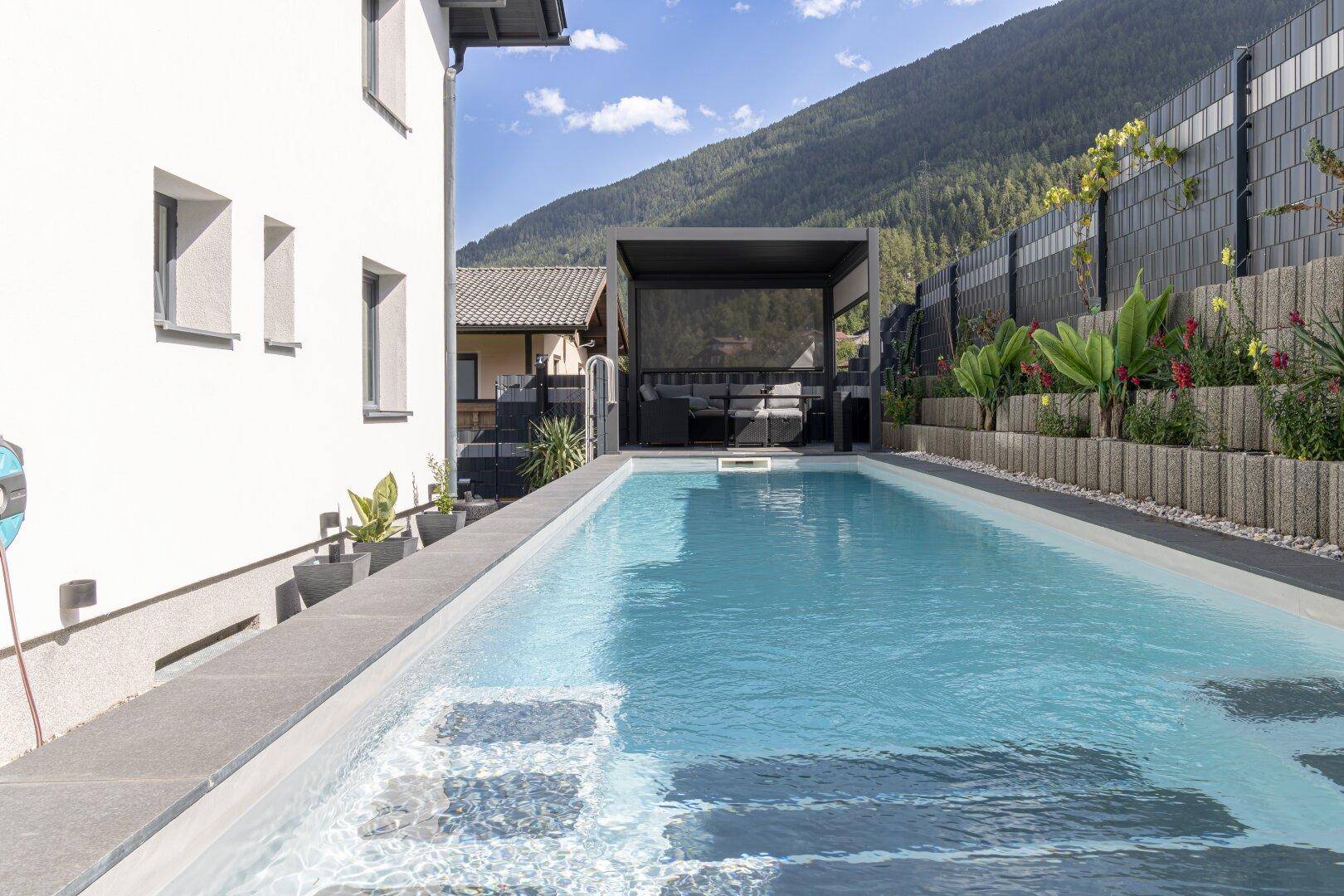 Langer, schmaler Außenpool neben dem Haus mit Blick auf die grünen Berge und eine Pergola im Hintergrund.