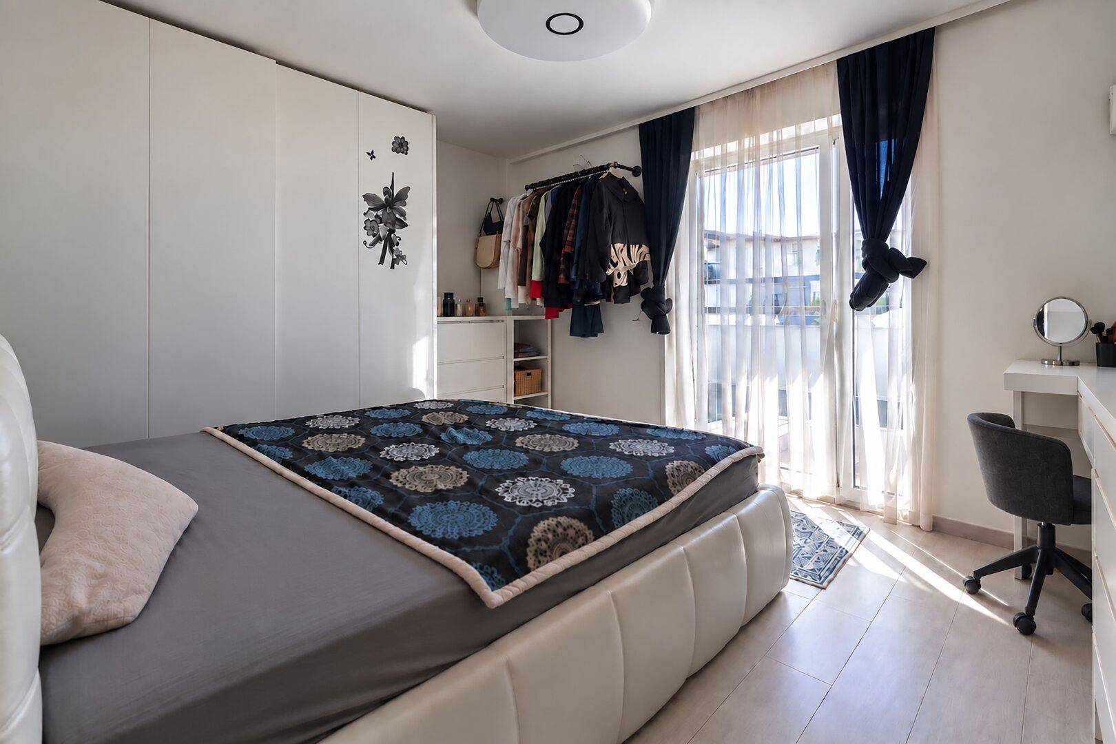 Helles Schlafzimmer mit Balkonzugang, weißem Kleiderschrank und einem Schreibtisch am Fenster.