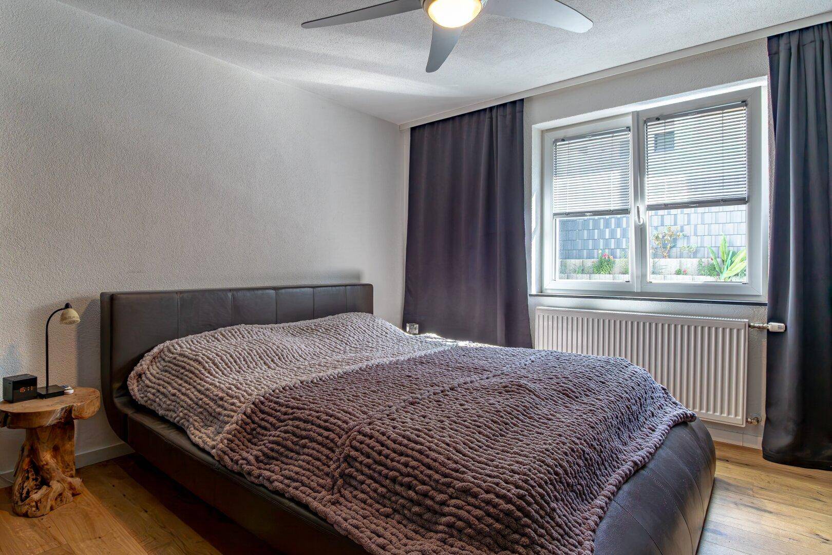 Schlafzimmer mit Doppelbett, Holzboden und einem Fenster mit Jalousien, gut beleuchtet.