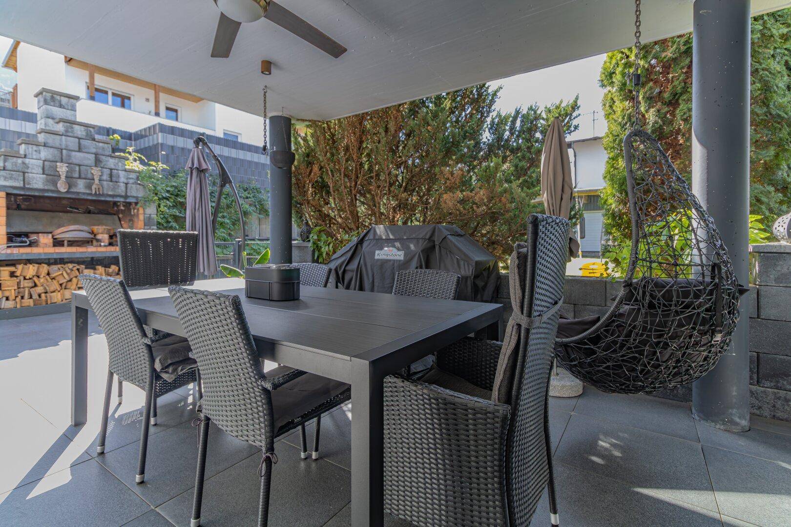 Überdachte Terrasse mit großem Esstisch, Grill und Hängesessel, umgeben von viel Grün.
