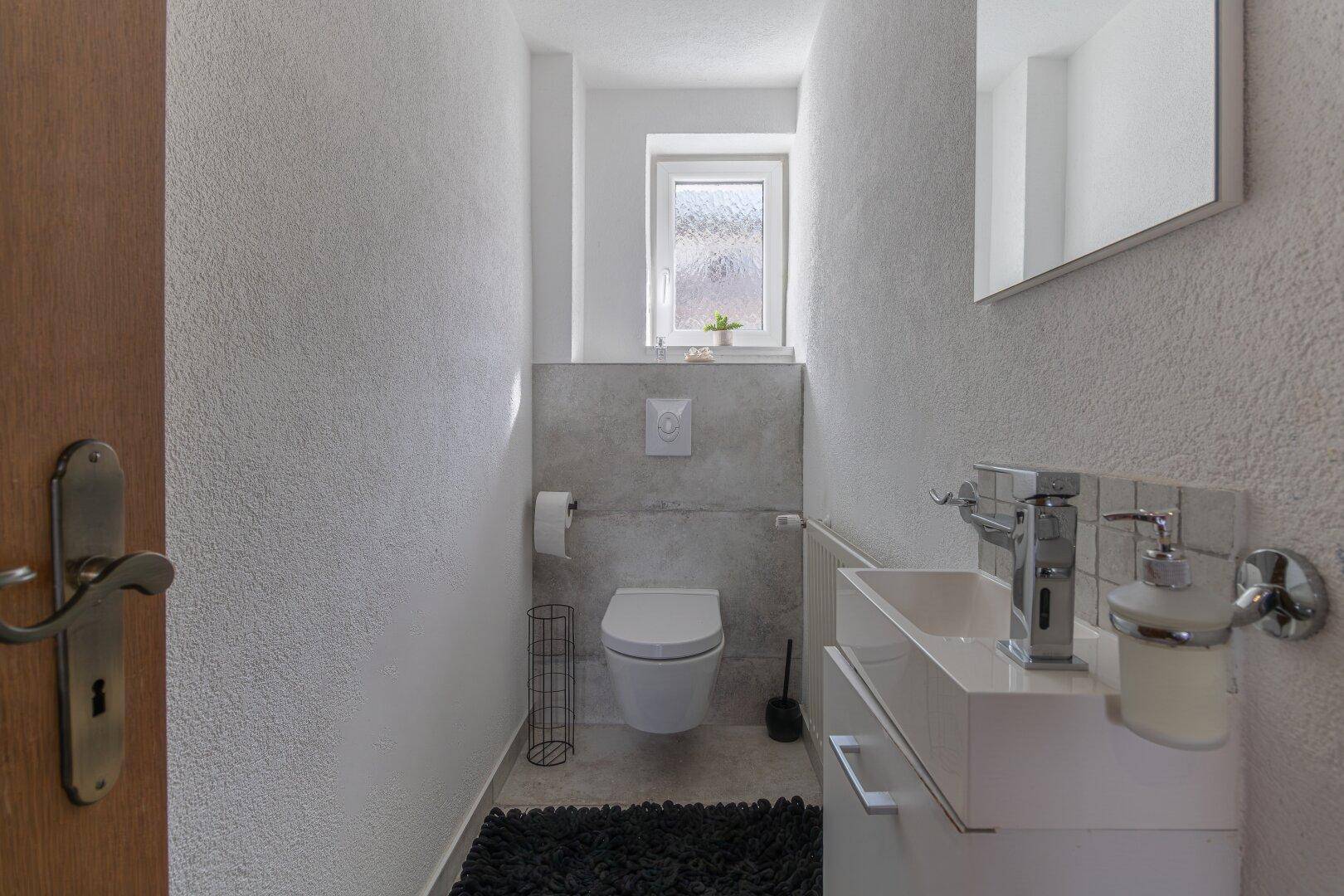 Modernes Gäste-WC mit wandhängendem WC, Waschbecken und einem kleinen Fenster für Tageslicht.