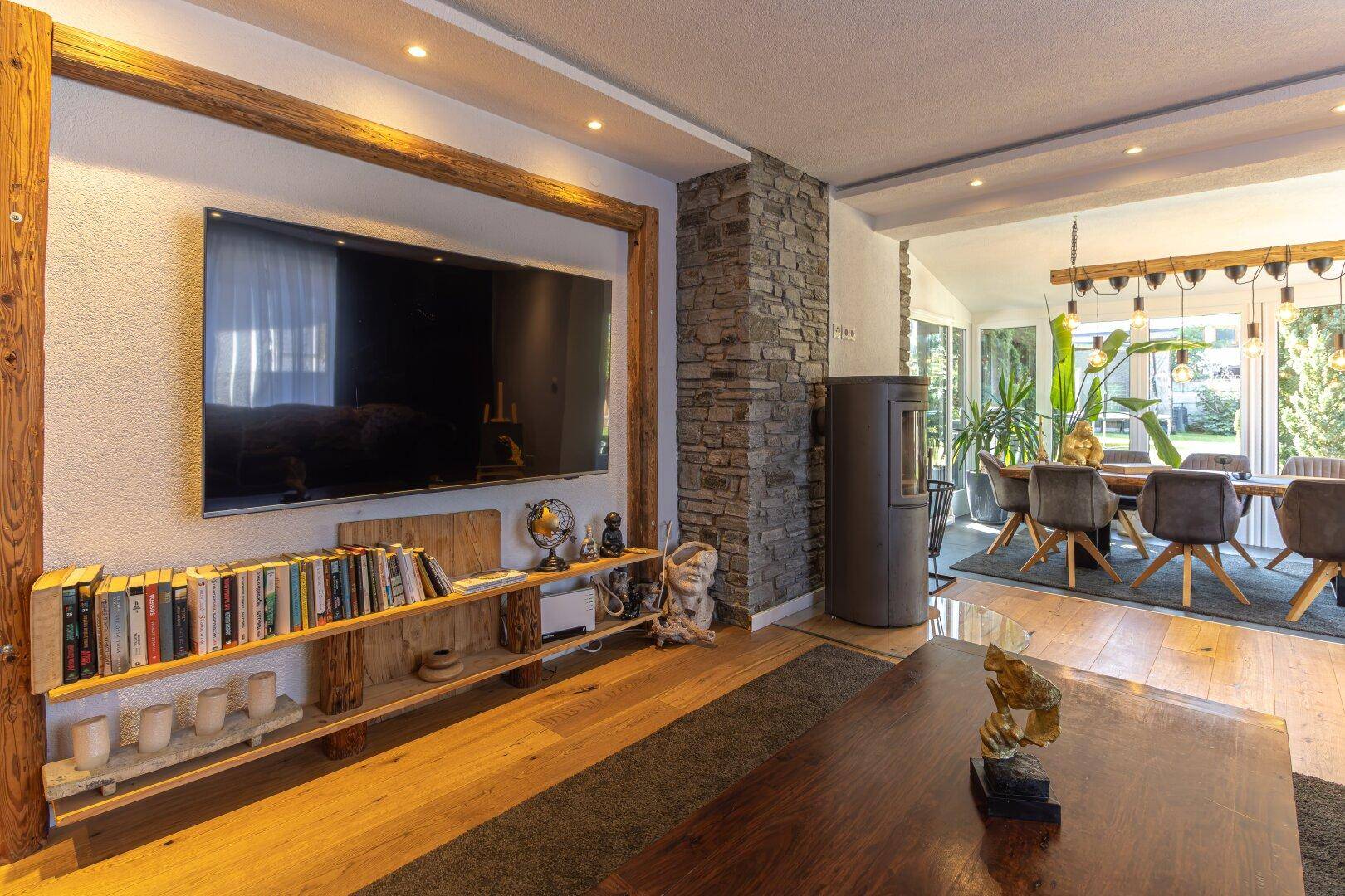 Gemütliches Wohnzimmer mit Holzboden, Kaminofen und großem Fernseher, offen zum Essbereich.