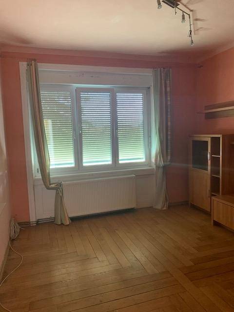 Gemütliches Wohnzimmer mit Holzparkettboden, Fenster mit Jalousien und einfacher Möblierung.